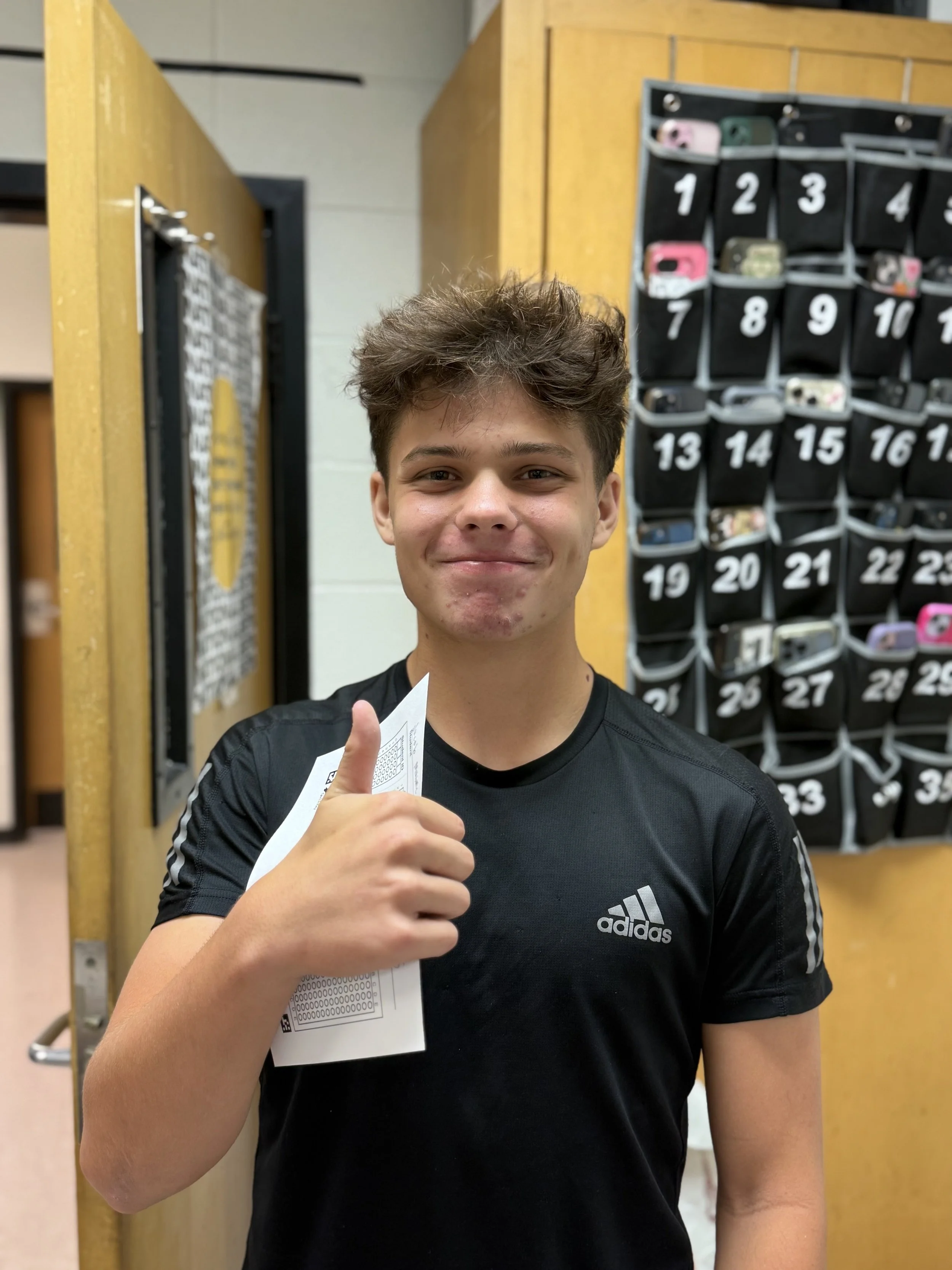 A young man with brown curly hair smiling and giving a thumbs-up, holding a piece of paper, in front of a wall-mounted organizer with numbered pockets, in an indoor setting.