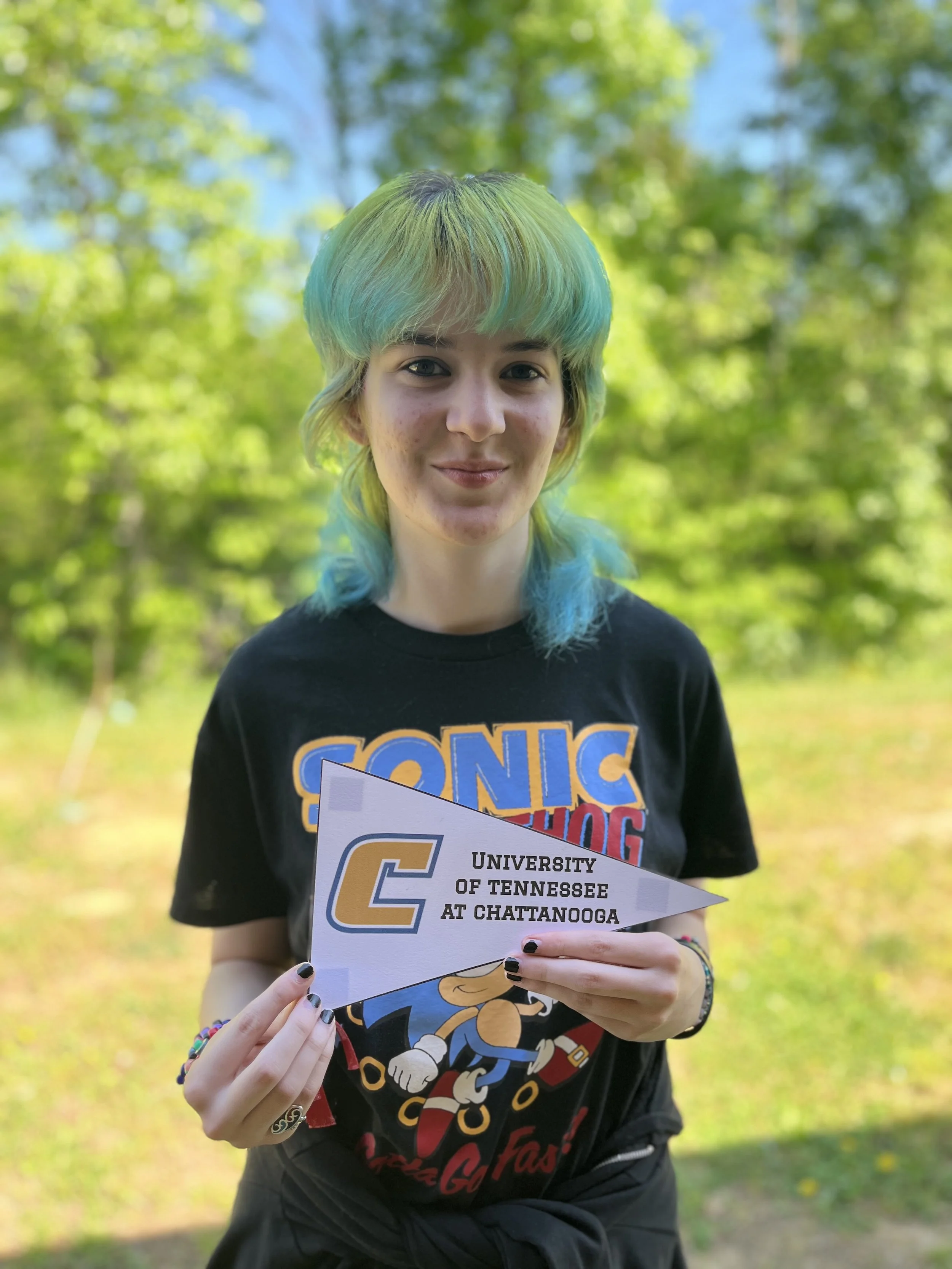 A young woman with bright pink, voluminous hair smiles at the camera. She has fair skin, gray eyes, and is wearing a black top with pink straps. She also has small earrings and a delicate necklace.