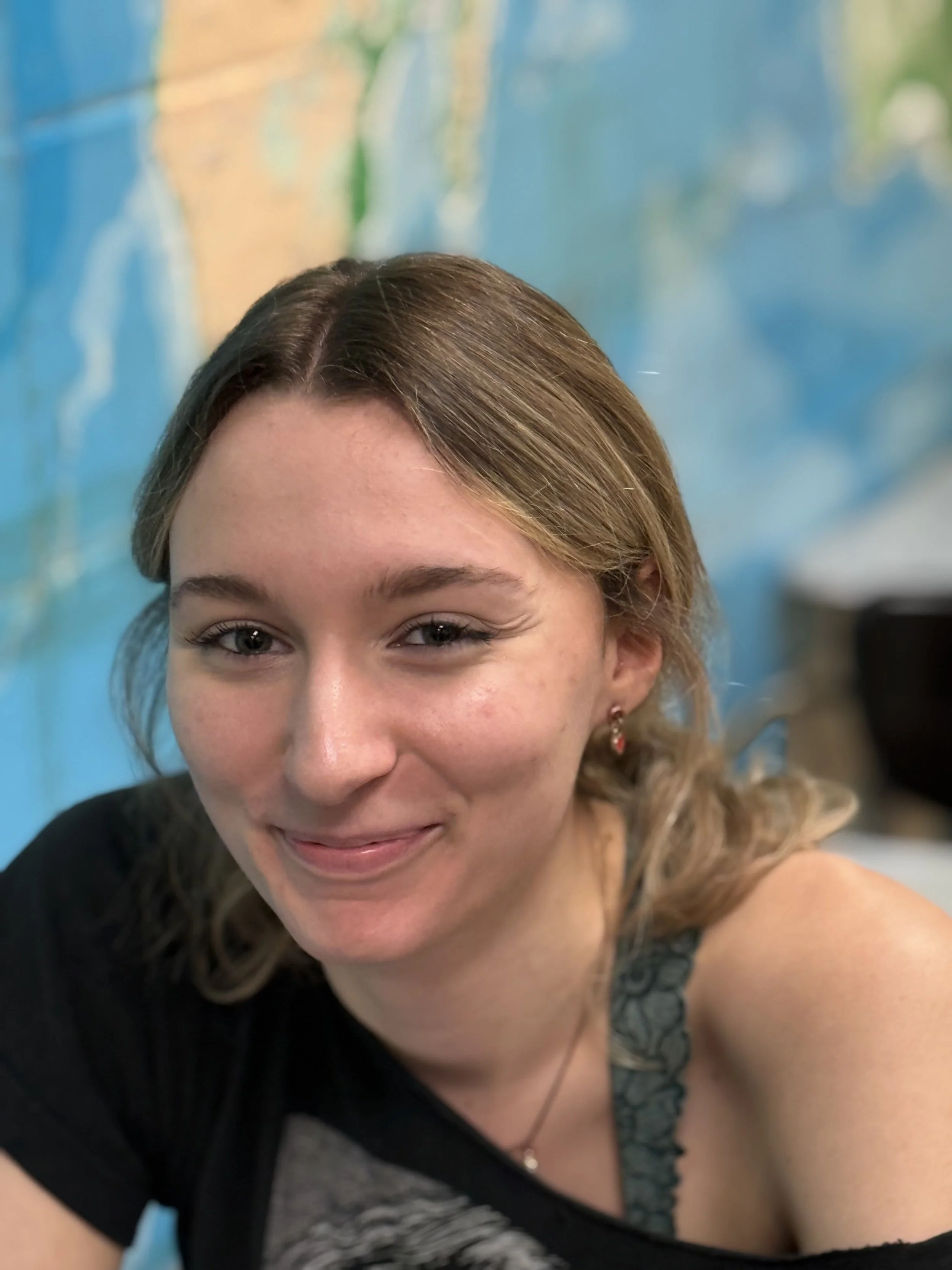 A young woman with shoulder-length light brown hair smiling, wearing a black top with lace shoulder detail, earrings, and a necklace, against a blurred colorful background.