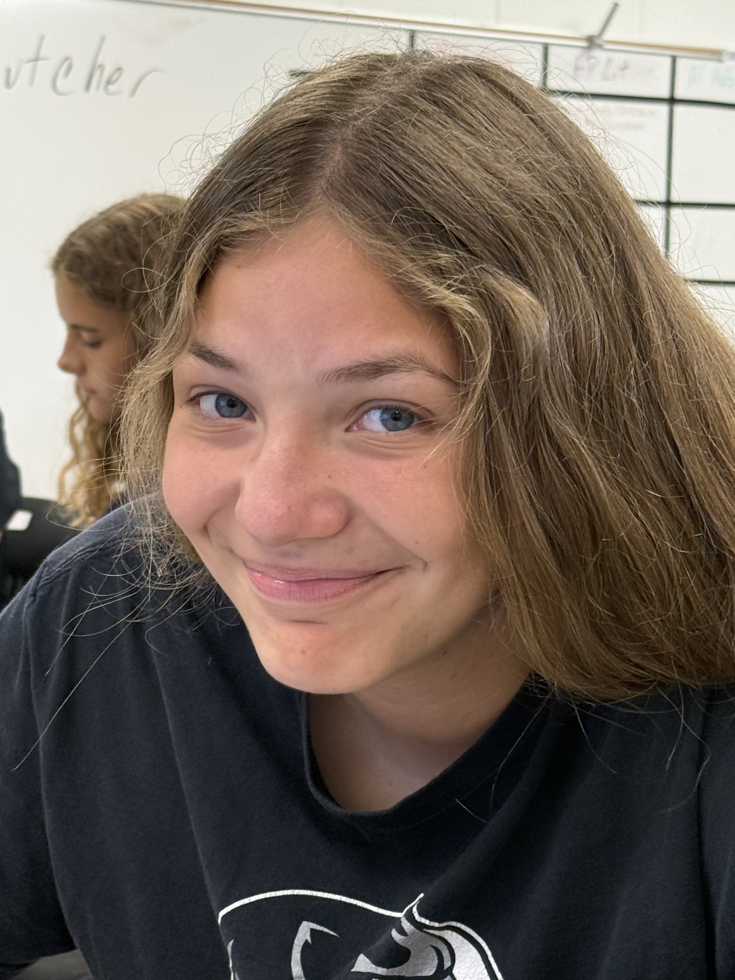 A young girl with long brown hair sitting at a desk in a classroom, smiling at the camera, with a notebook open with colorful highlights in front of her. There is a map on the wall behind her.
