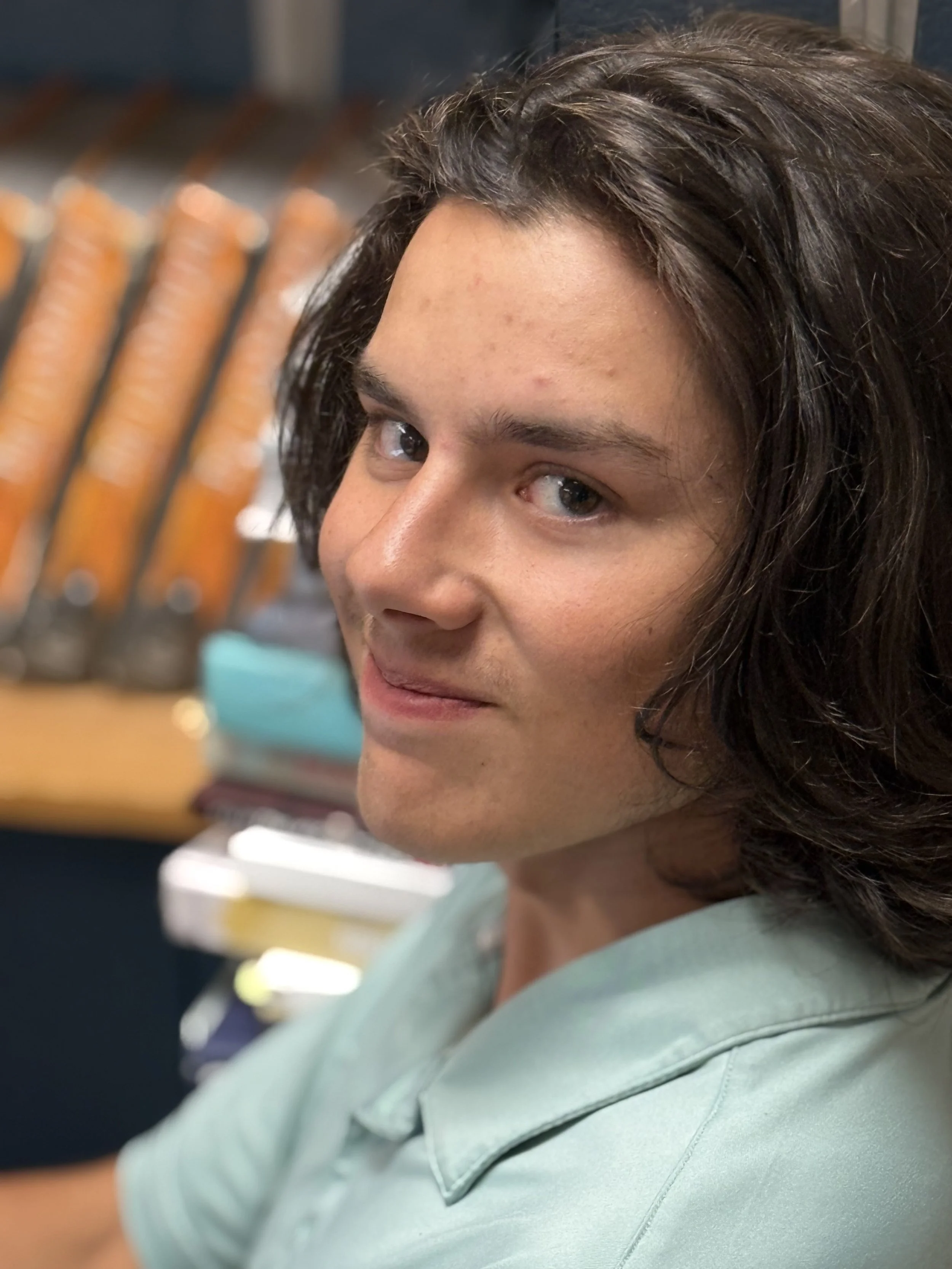 Close-up of a young man with shoulder-length dark hair, blue eyes, and light skin, wearing a light green polo shirt, with a blurred background of knives in a knife block.