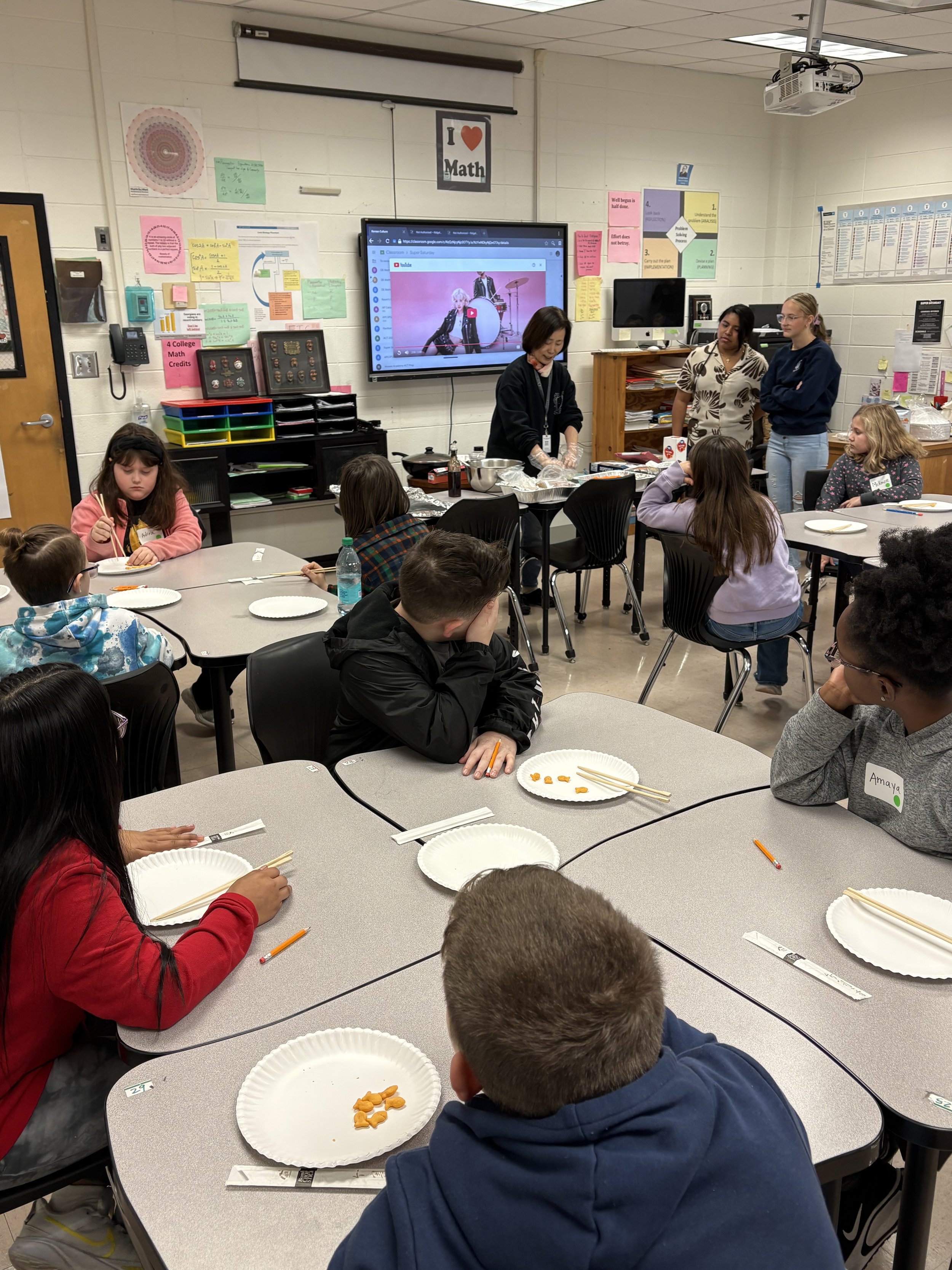 A classroom with students seated at tables, some with plates and chopsticks, watching a woman demonstrate a cooking activity at the front. There is a large screen displaying a YouTube video, colorful posters on the walls, and a teacher or instructor 