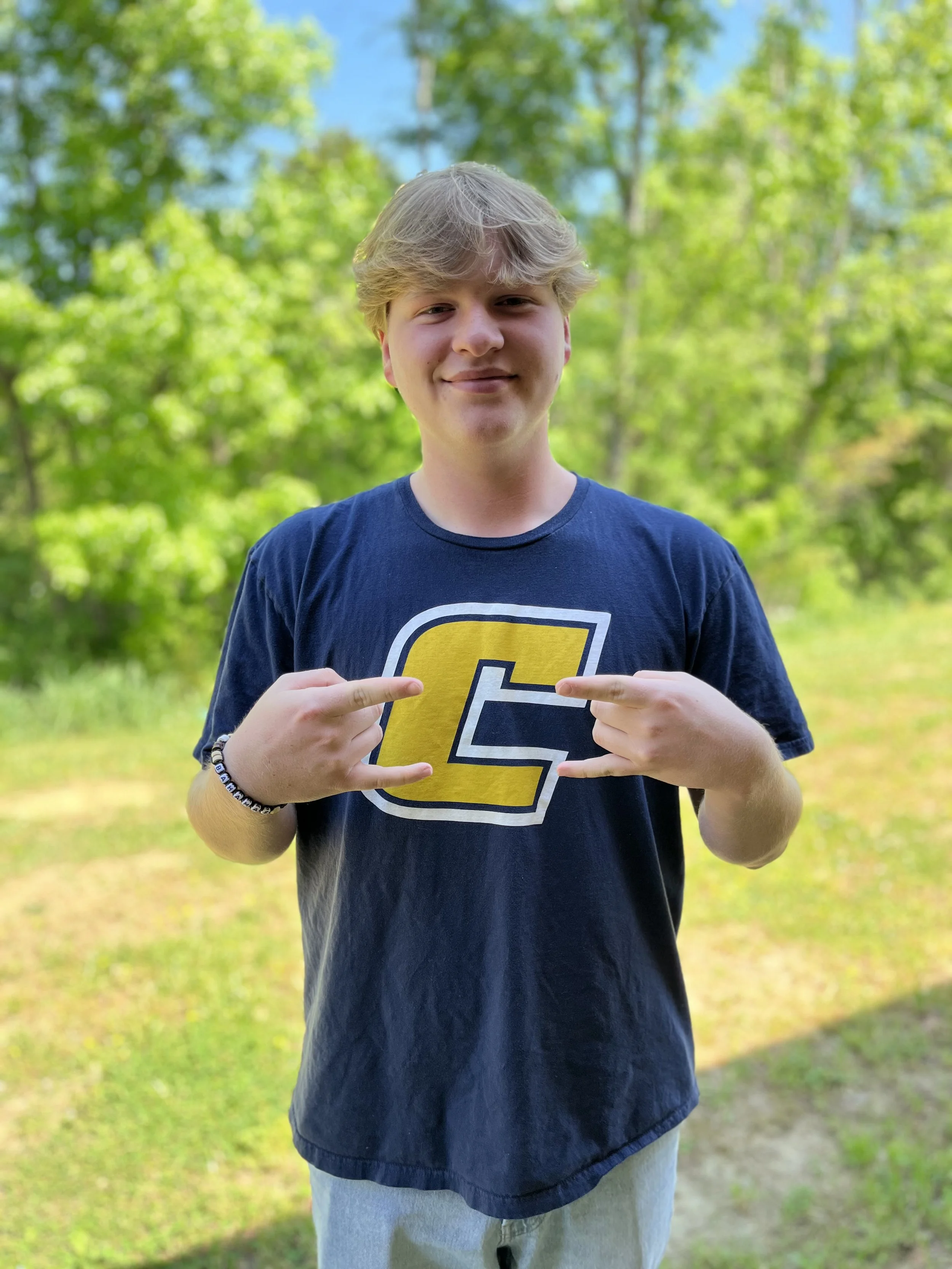 A young man with light brown, wavy hair smiling at the camera. He is wearing a beige polo shirt. Background shows bookshelves with red and yellow books, and another person partially visible behind him.