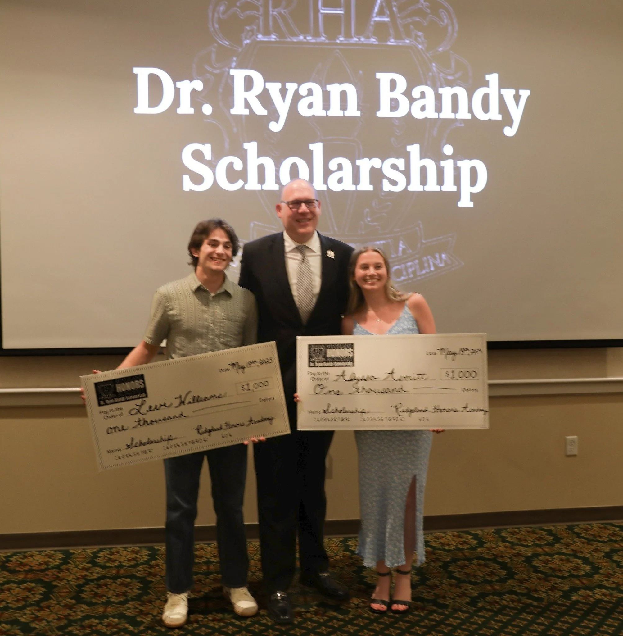 Three people standing in front of a screen displaying 'Dr. Ryan Bandy Scholarship'. The person in the middle is holding a large check for $1,000 made out to Lewis Hallam. The person on the right is holding a check for $1,000 made out to Aurora Amonte. All three are smiling.