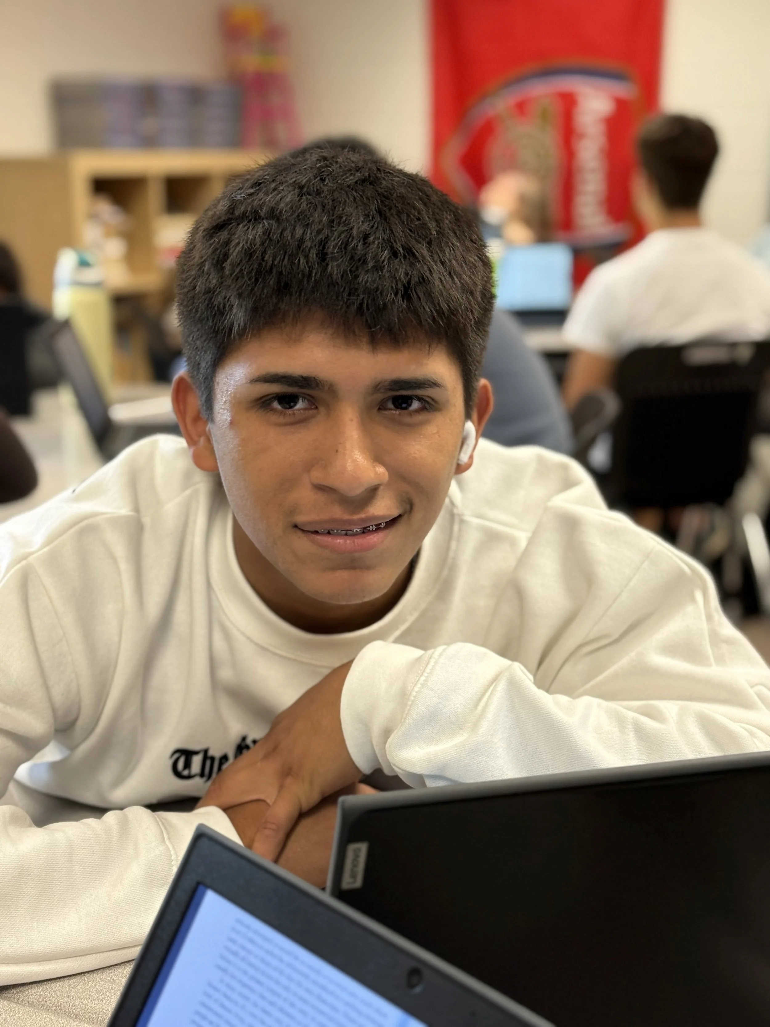 A teenage boy with short dark hair smiling at the camera in a classroom, wearing a white sweatshirt, with students and computers in the background.