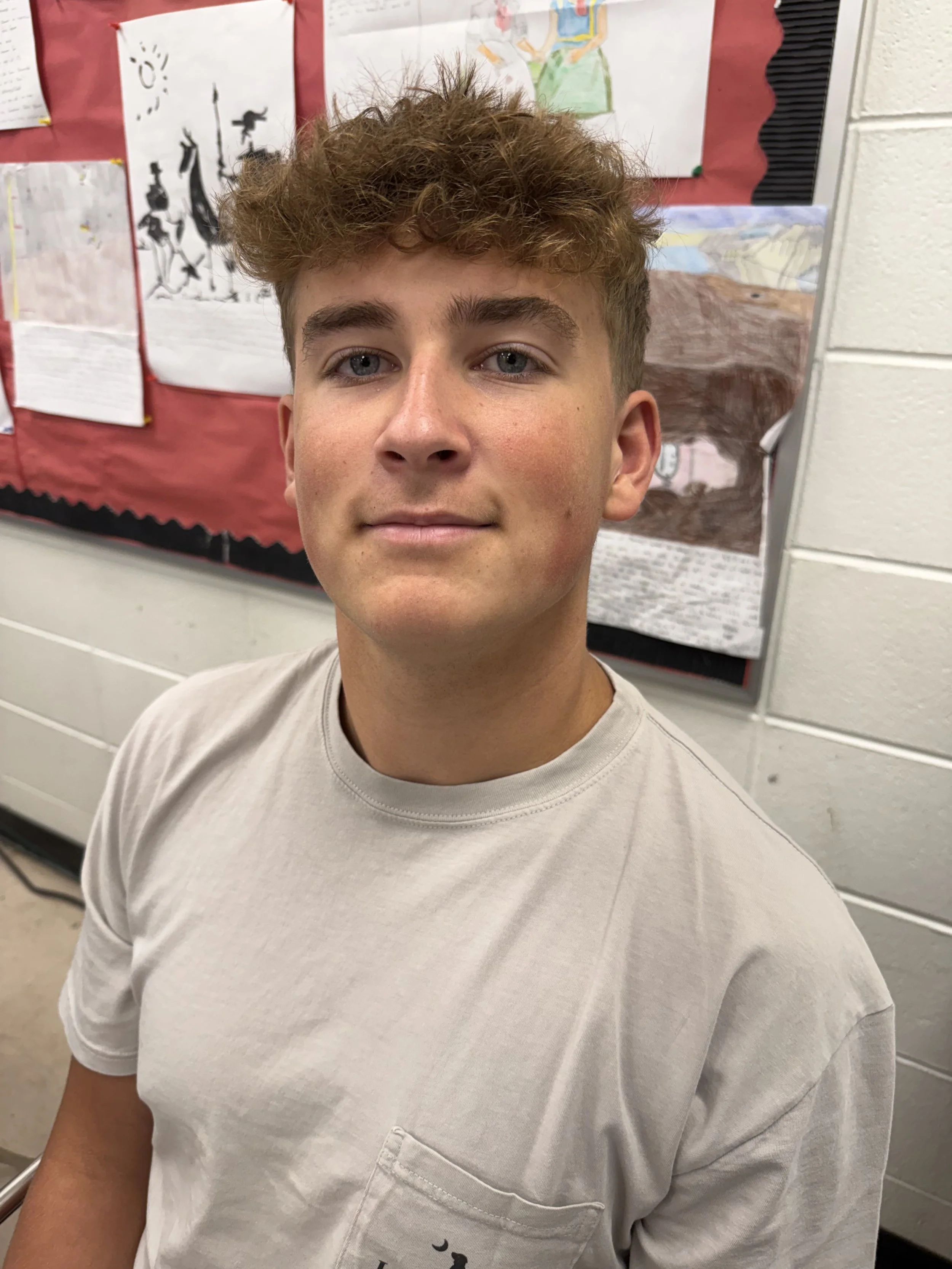 A young man with curly light brown hair and blue eyes, wearing a black shirt, looking at the camera with a slight smile inside a busy indoor setting.