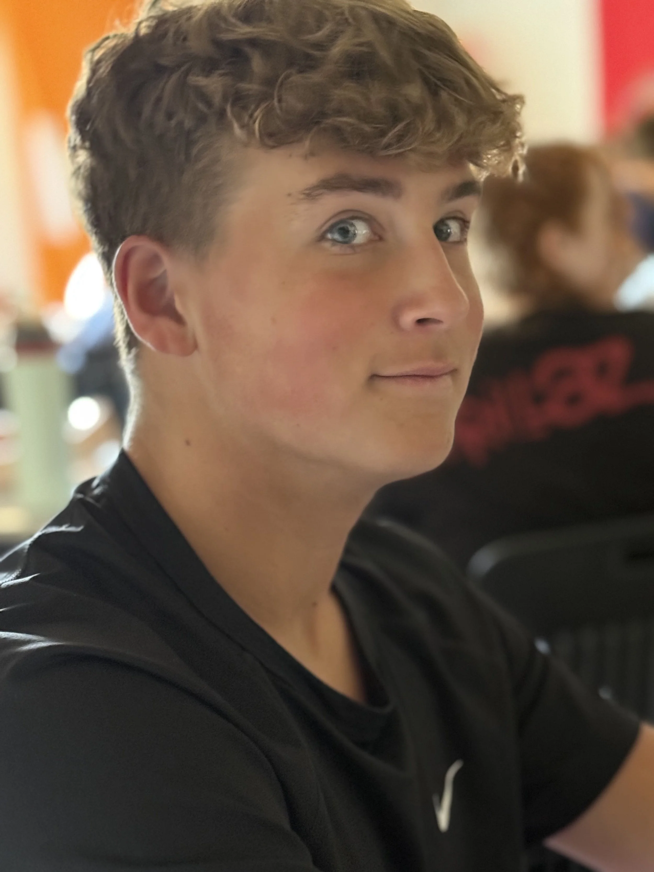 A young man with curly light brown hair and blue eyes, wearing a black shirt, looking at the camera with a slight smile inside a busy indoor setting.