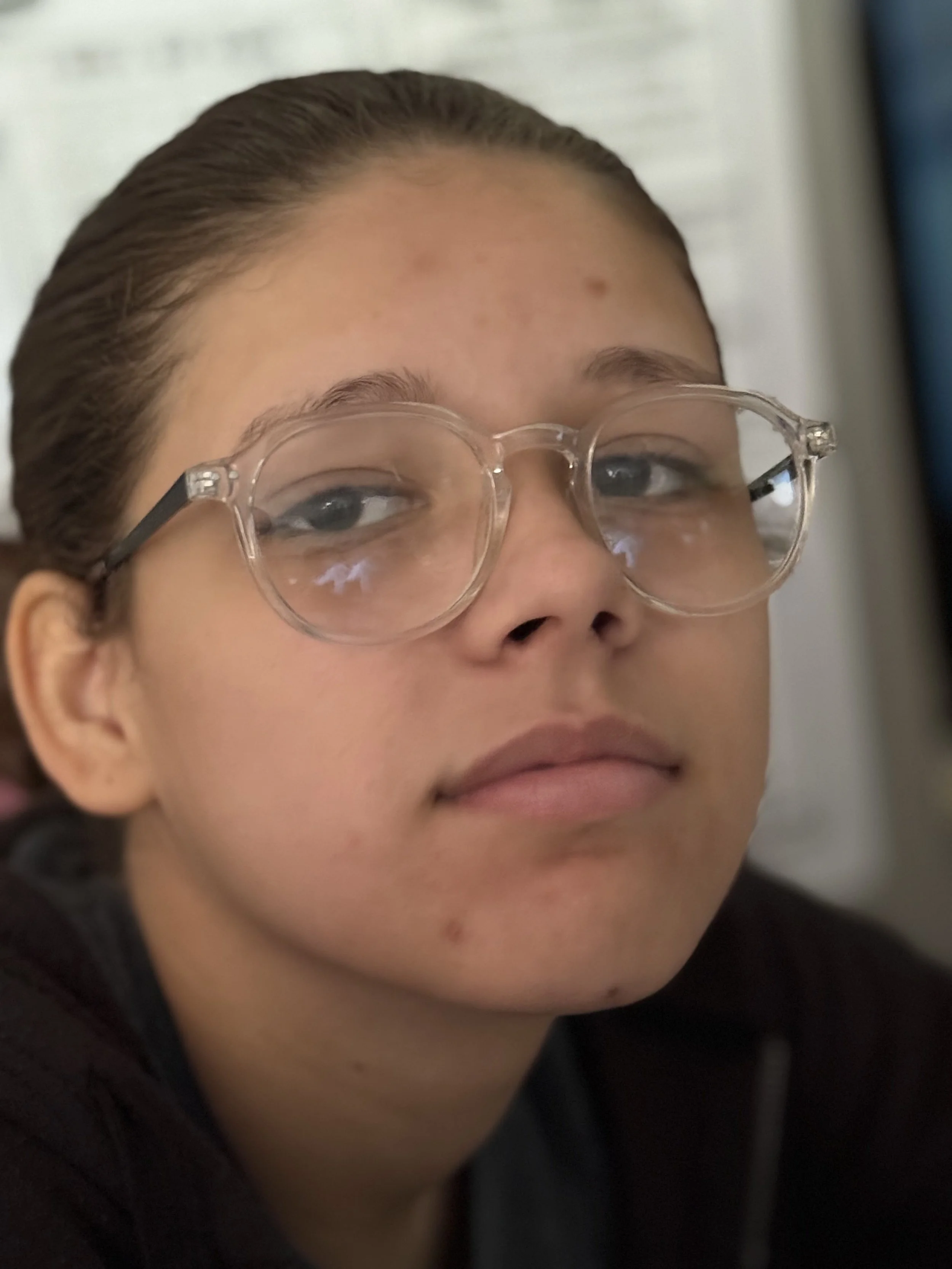 Close-up of a young person's face wearing clear glasses, with brown hair slicked back and a slight smile.