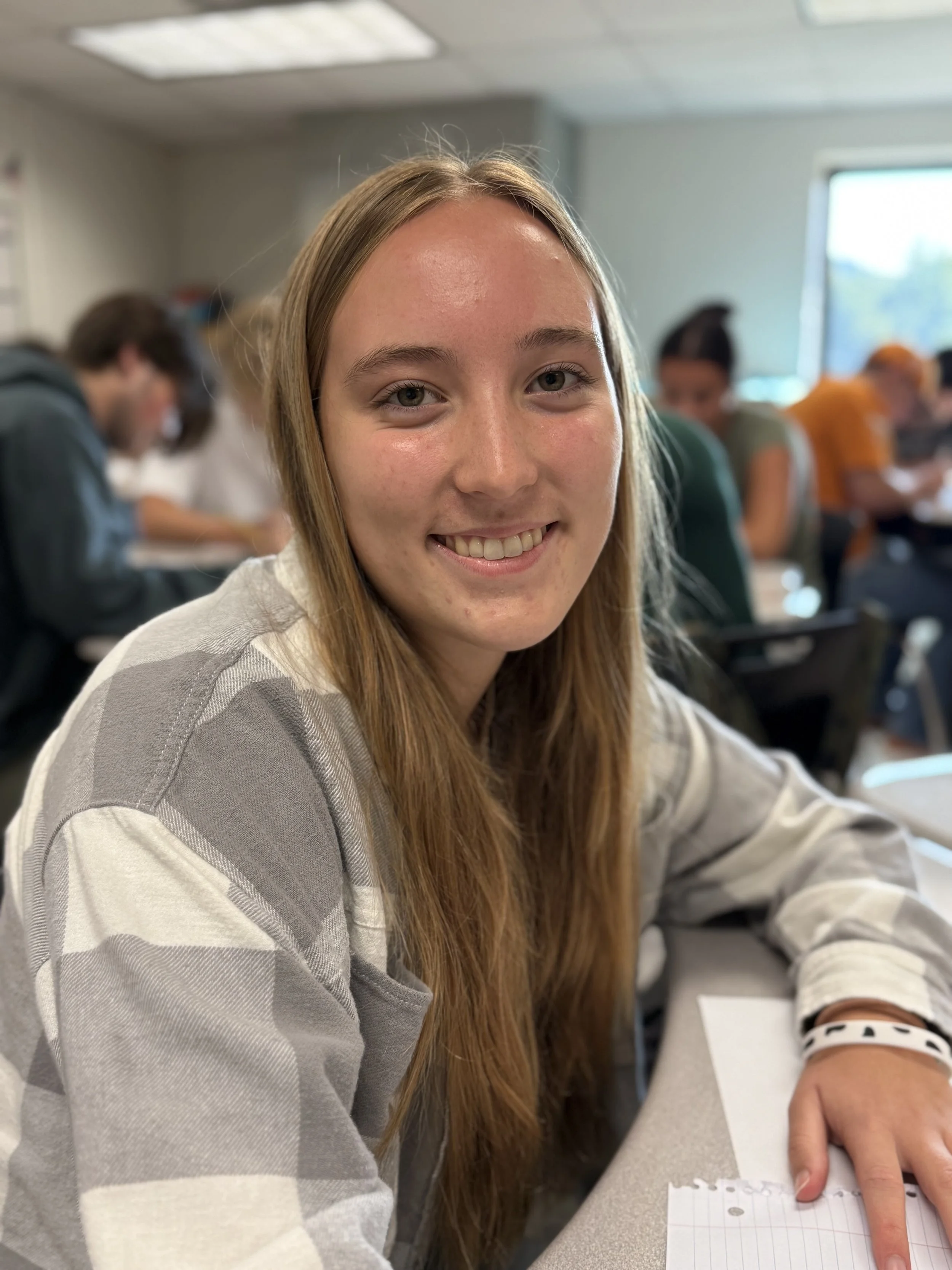 Young girl with long red hair smiling at camera in classroom setting with students in background.