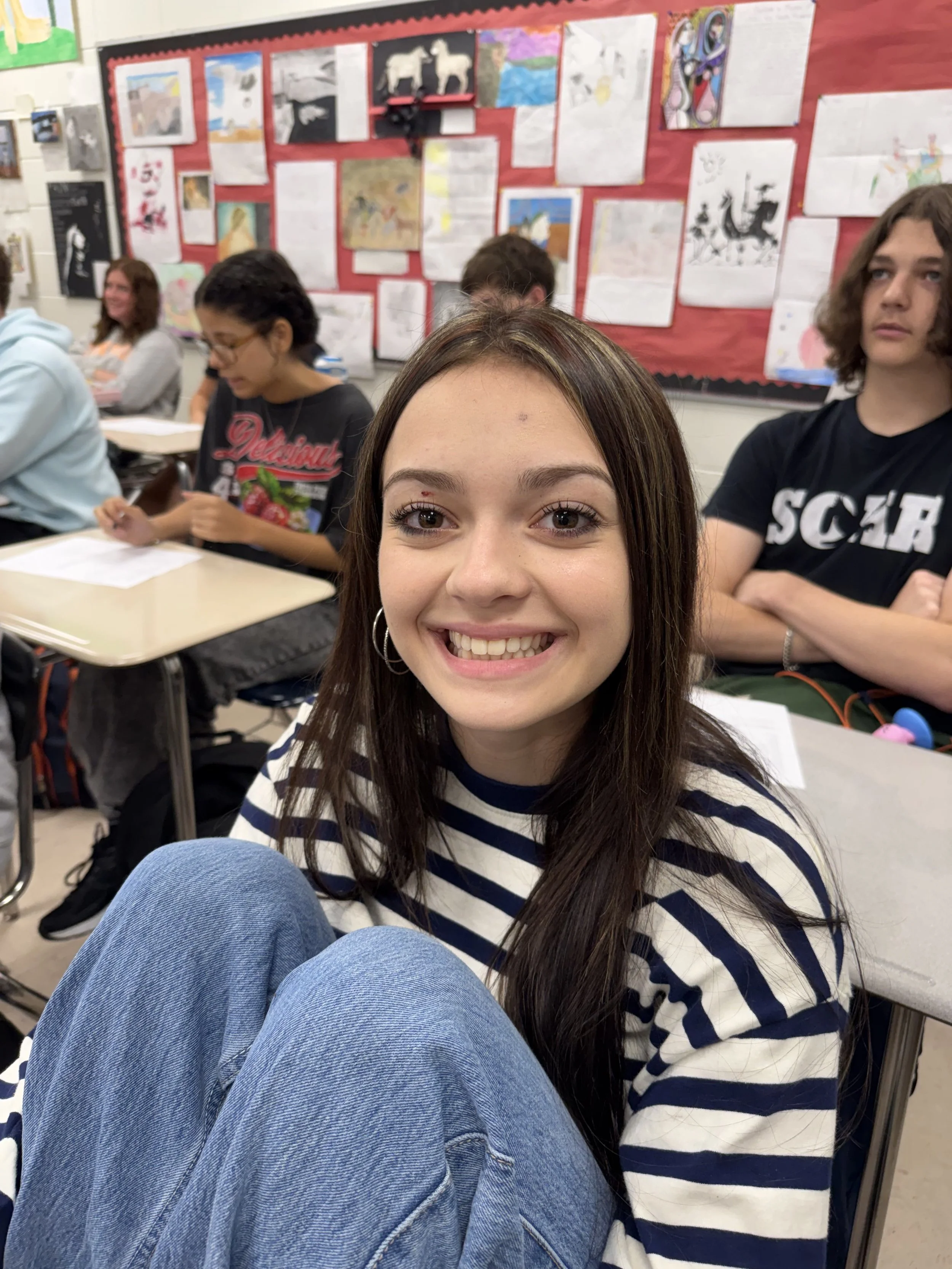 A young girl with long brown hair and a white shirt, sitting cross-legged at a desk in a classroom, smiling at the camera.