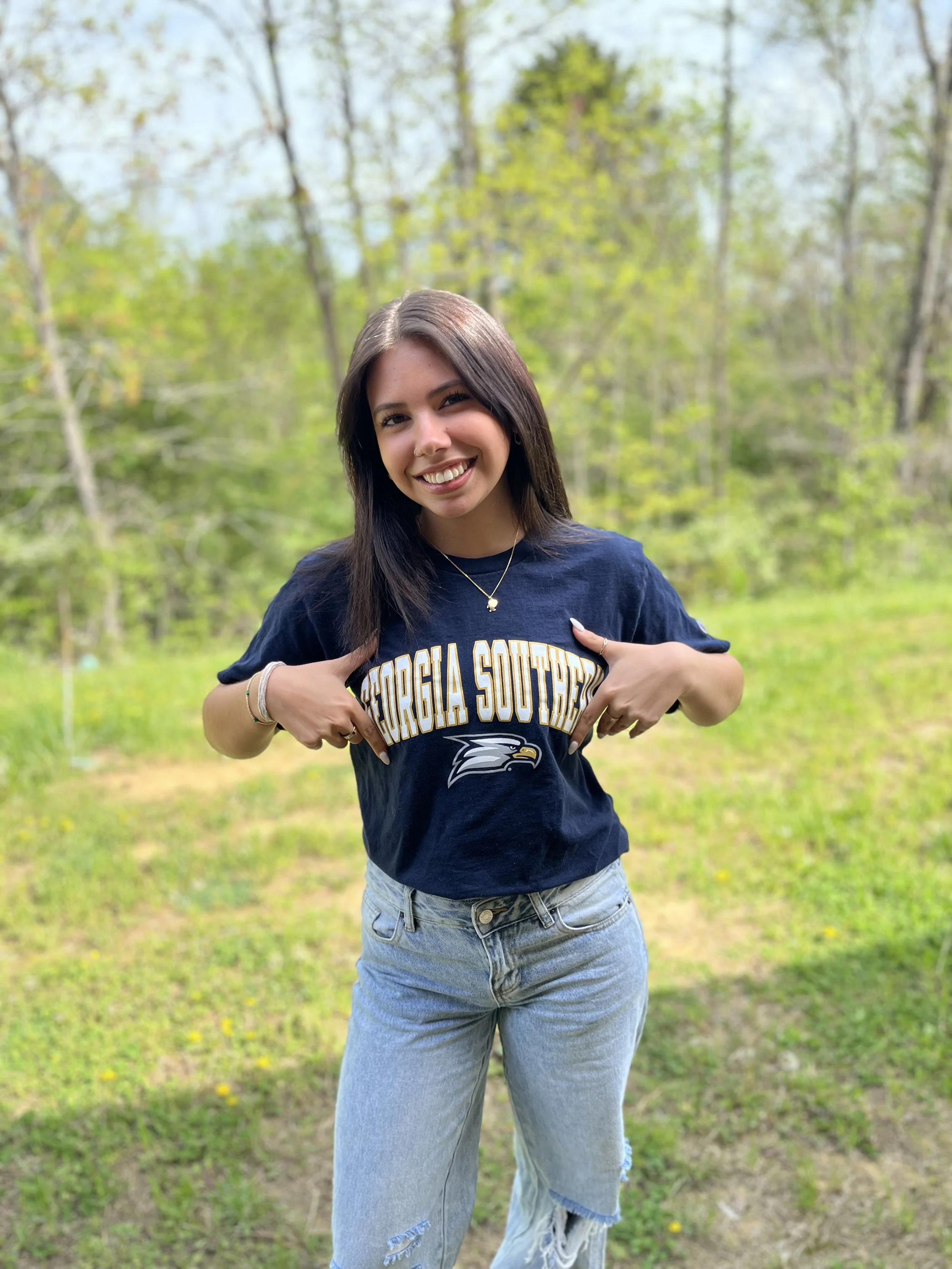 A young woman with dark brown hair, brown eyes, and a nose ring smiling at the camera, wearing a leopard-print top and layered necklaces, with a white cardigan draped over her shoulders.