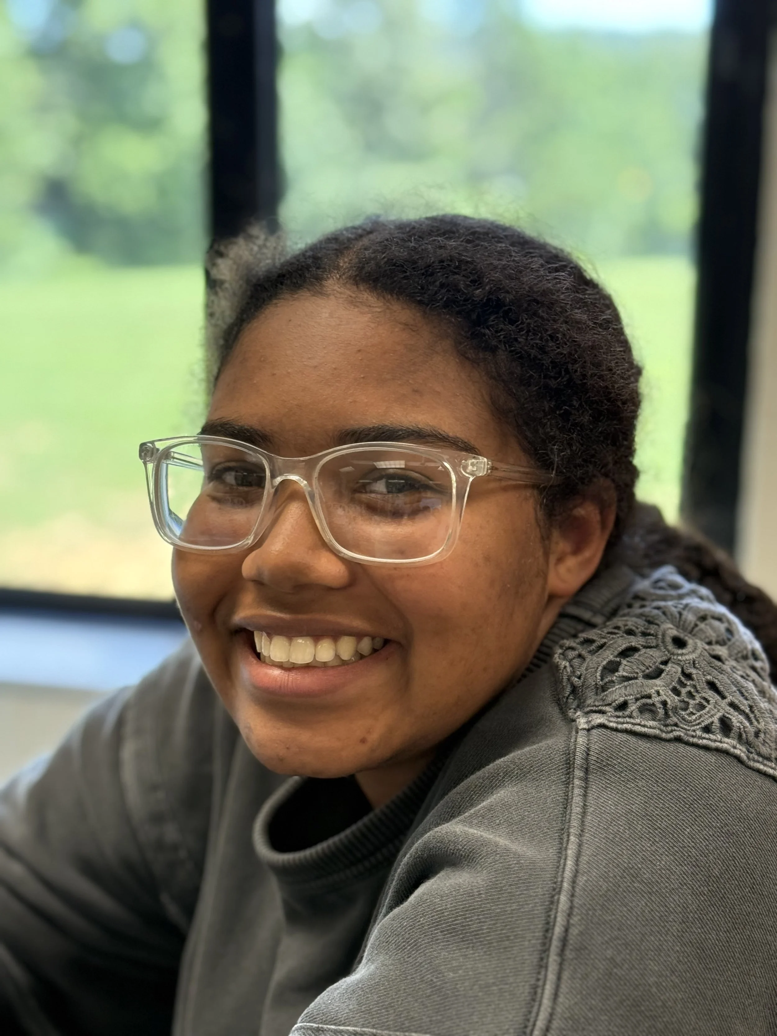 A young woman with glasses smiling, sitting indoors near large windows with a green outdoor landscape visible in the background.
