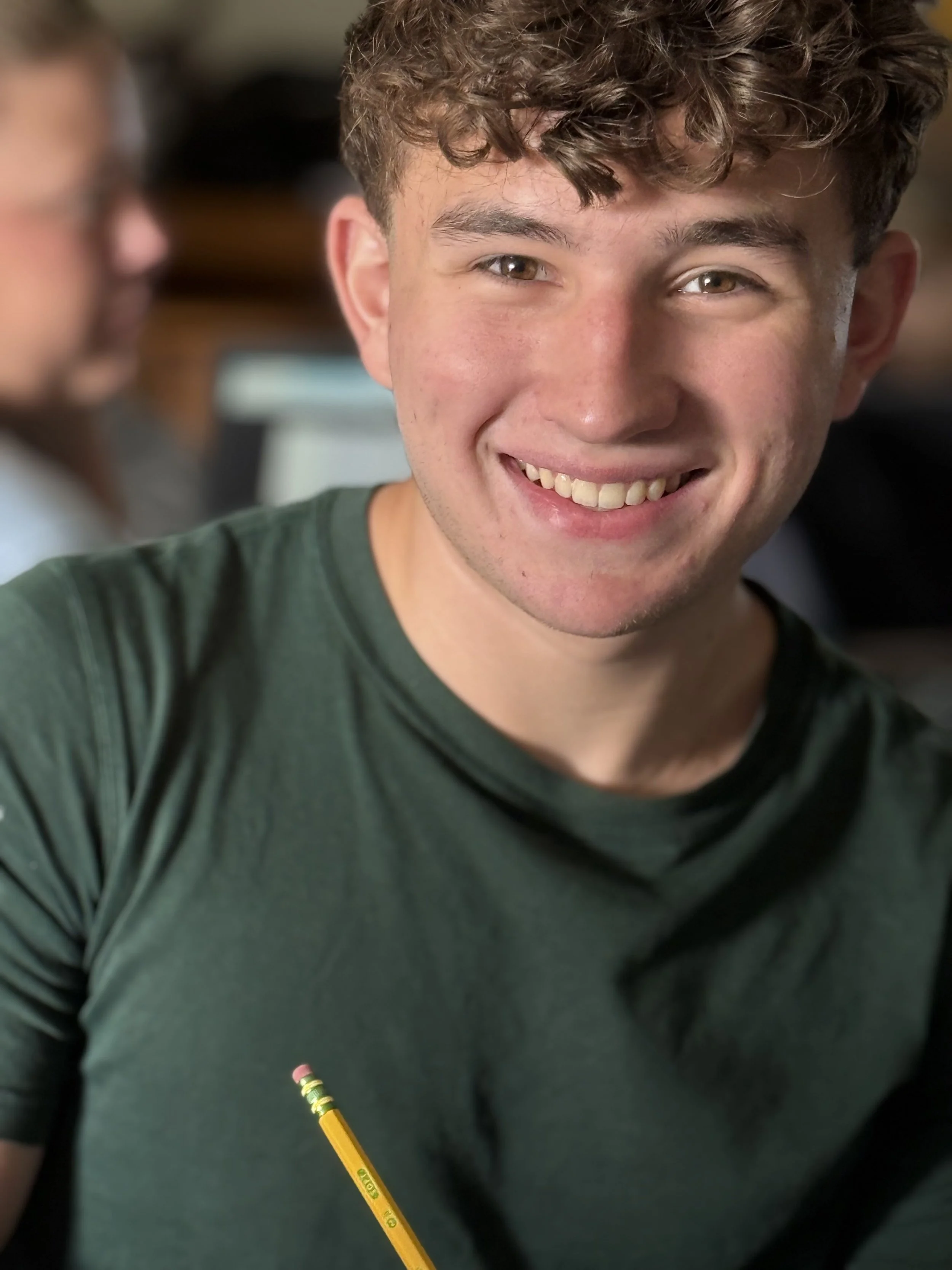 A young man with curly brown hair and a bright smile, wearing a dark green shirt, sitting with a yellow pencil in front of him. In the background, there is a woman with glasses working on a laptop.