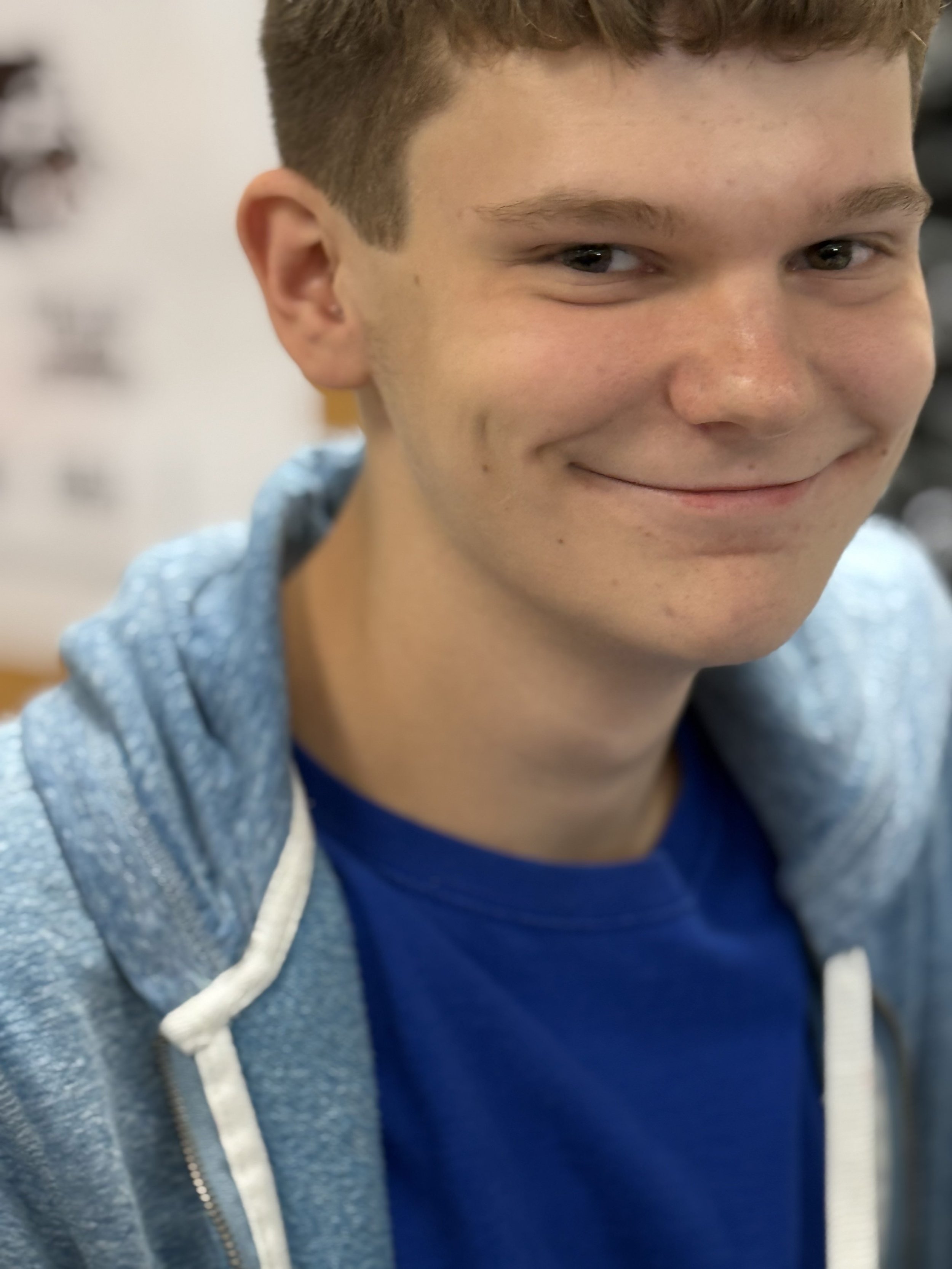 Close-up of a teenage boy smiling, wearing a blue T-shirt and a light blue hoodie, indoors.