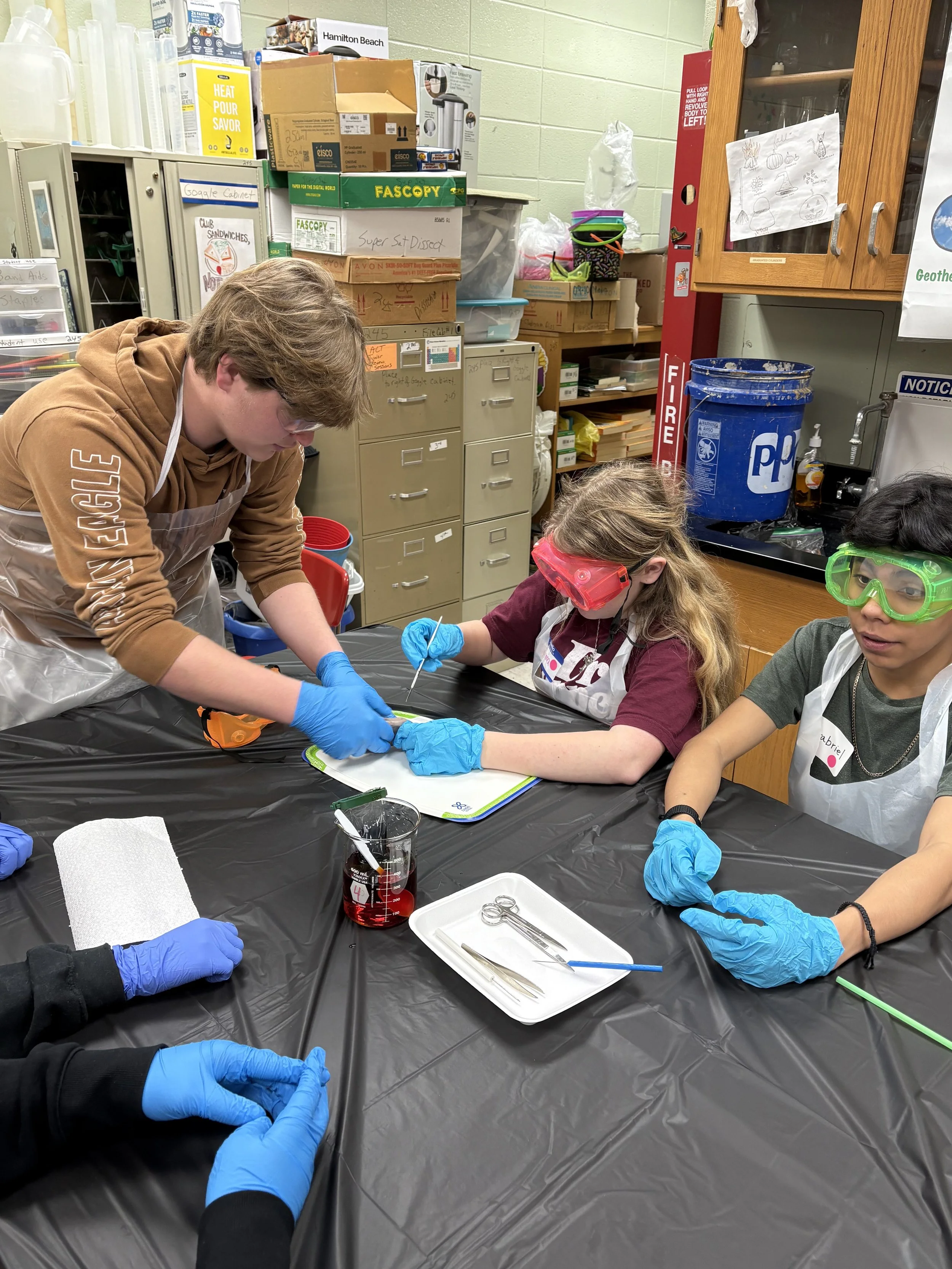 Three children wearing safety goggles and gloves are seated at a black table in a science classroom, conducting an experiment with a teacher guiding them. The background features shelves and cabinets with various supplies, and the children are focuse