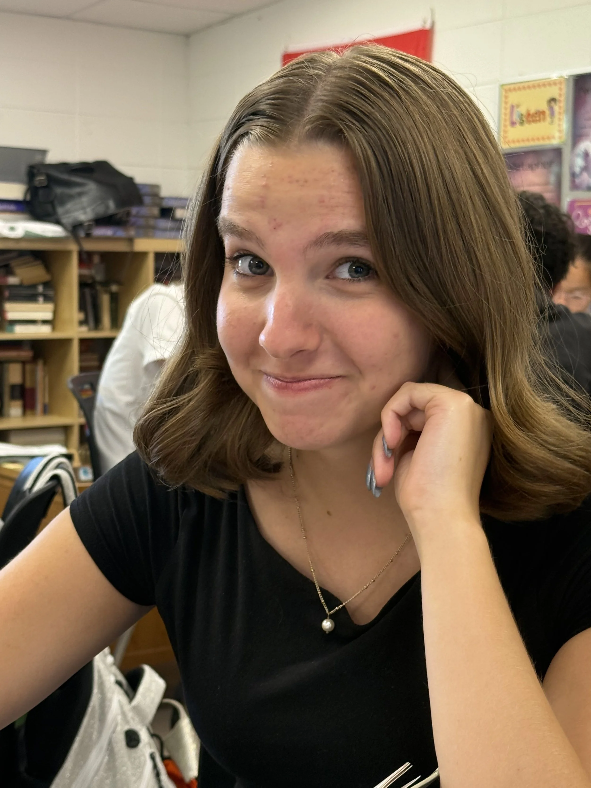Young woman with brown wavy hair sitting at a desk with a laptop, smiling, resting her chin on her hand, in a classroom or office setting.