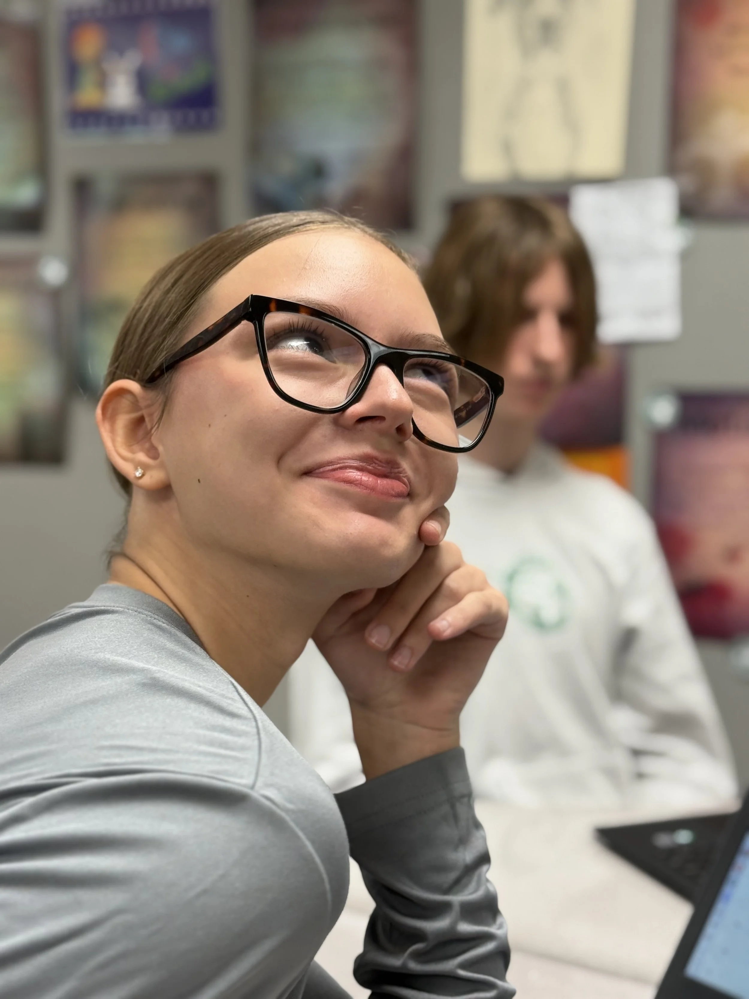 A young woman with glasses smiling and resting her chin on her hand, seated in a classroom with a young man in the background, and posters on the wall.