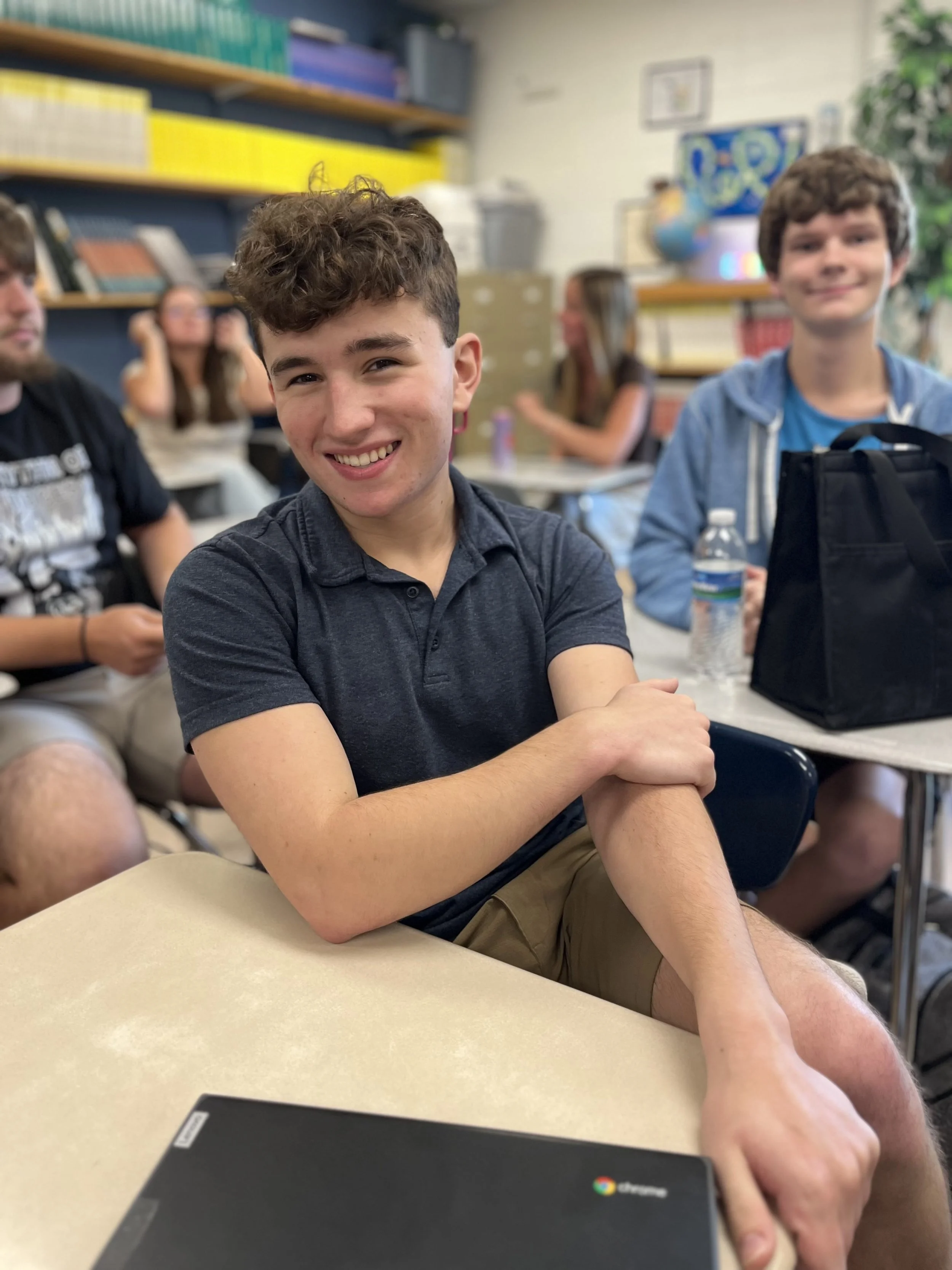 A young man with curly brown hair and a bright smile, wearing a dark green shirt, sitting with a yellow pencil in front of him. In the background, there is a woman with glasses working on a laptop.