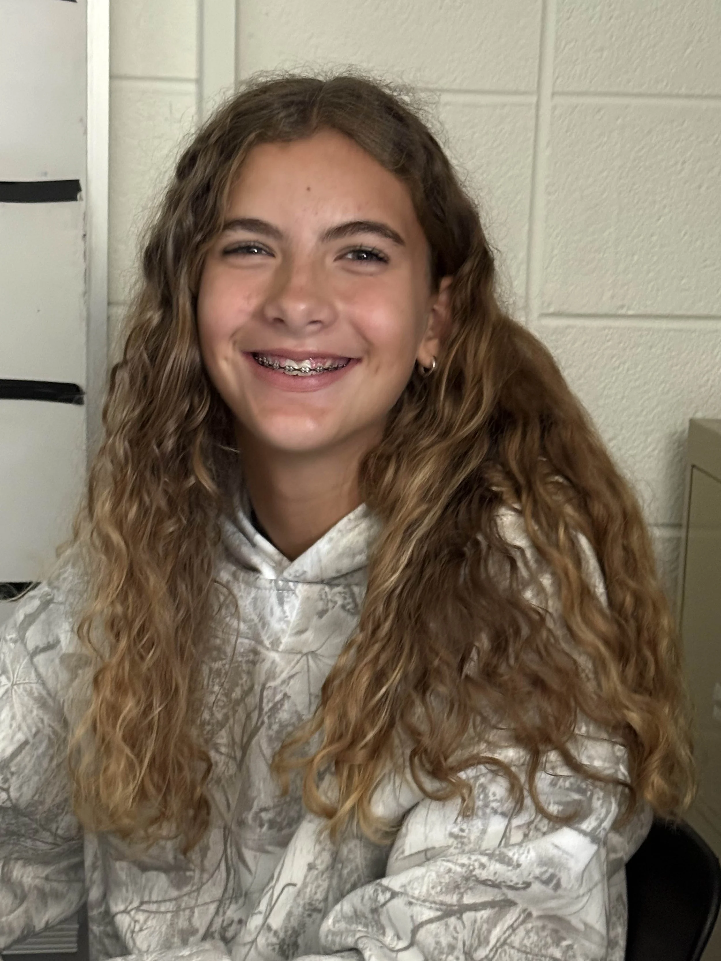 A young girl with curly, long light brown hair smiling at the camera, wearing braces and small earrings, in a room with beige walls and a filing cabinet.