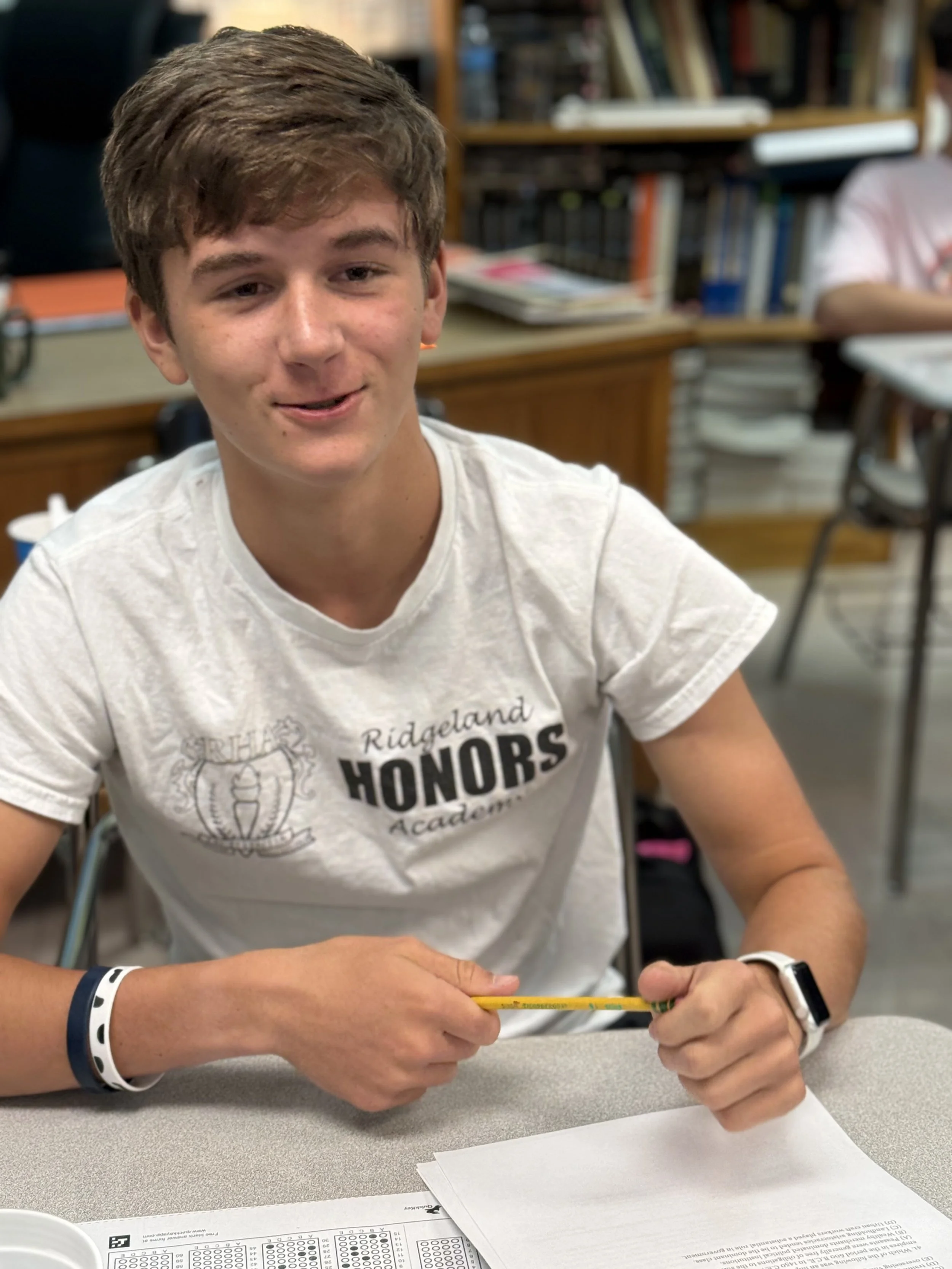 A young man sitting at a classroom desk, holding a yellow pencil, with papers in front of him, wearing a white t-shirt that says "Ridge Land Honors Academy" and a smartwatch on his left wrist.