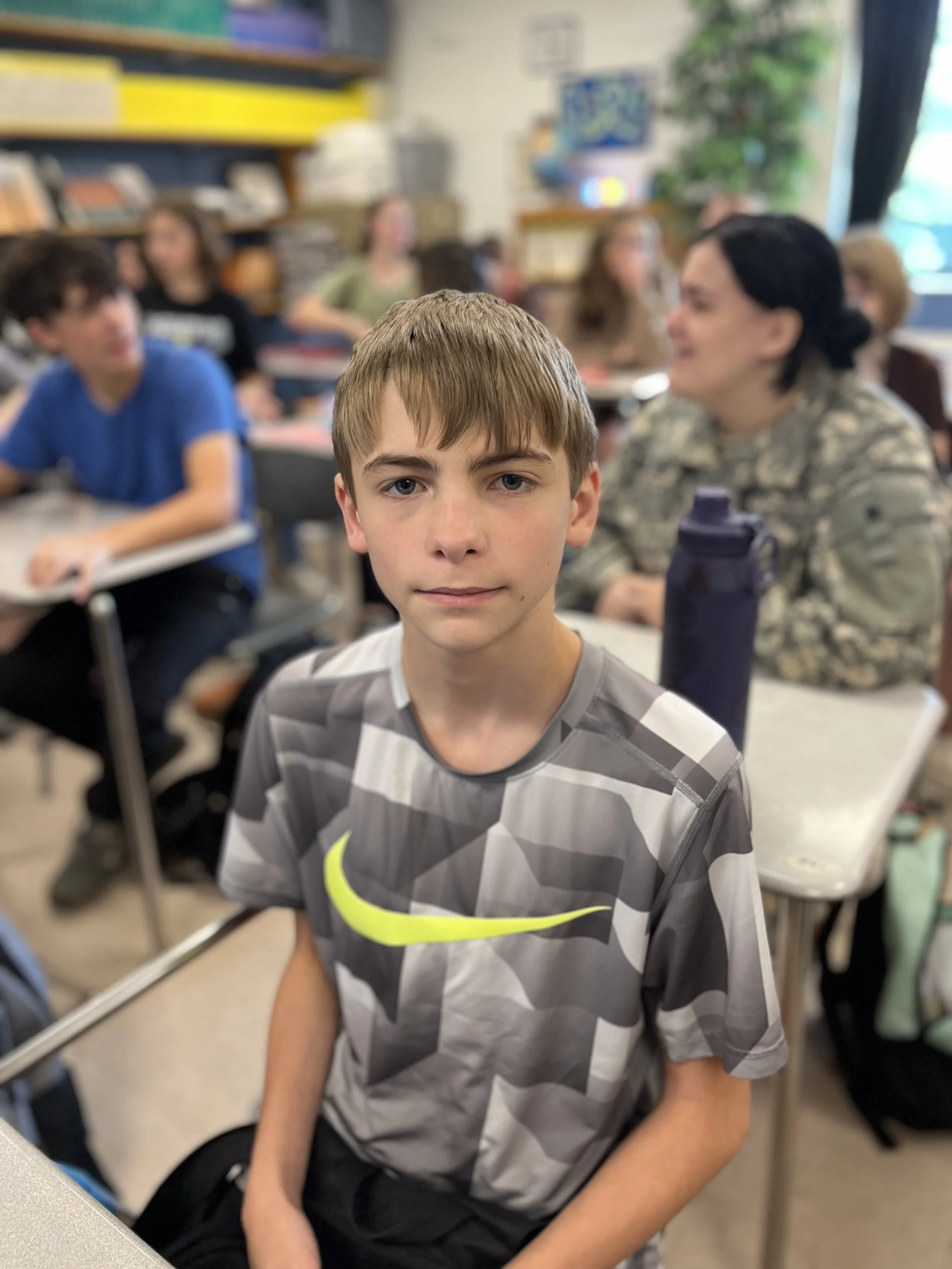 A young boy with brown hair and wearing a blue shirt looks up at the camera in a classroom, sitting at a desk with a green notebook and a pencil in front of him. Other students are visible in the background.