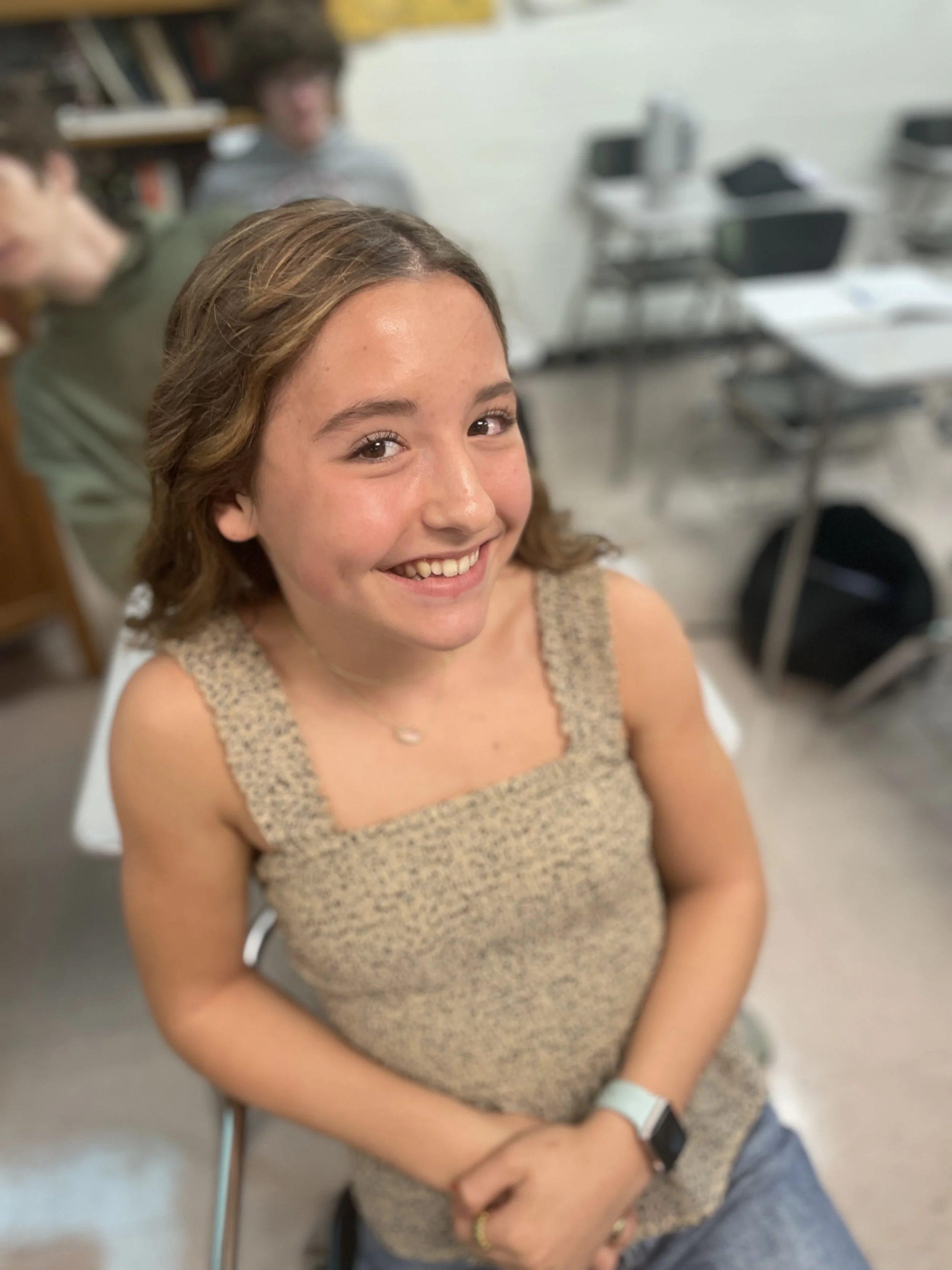 A young woman smiling, sitting in a classroom, with two other students blurred in the background.