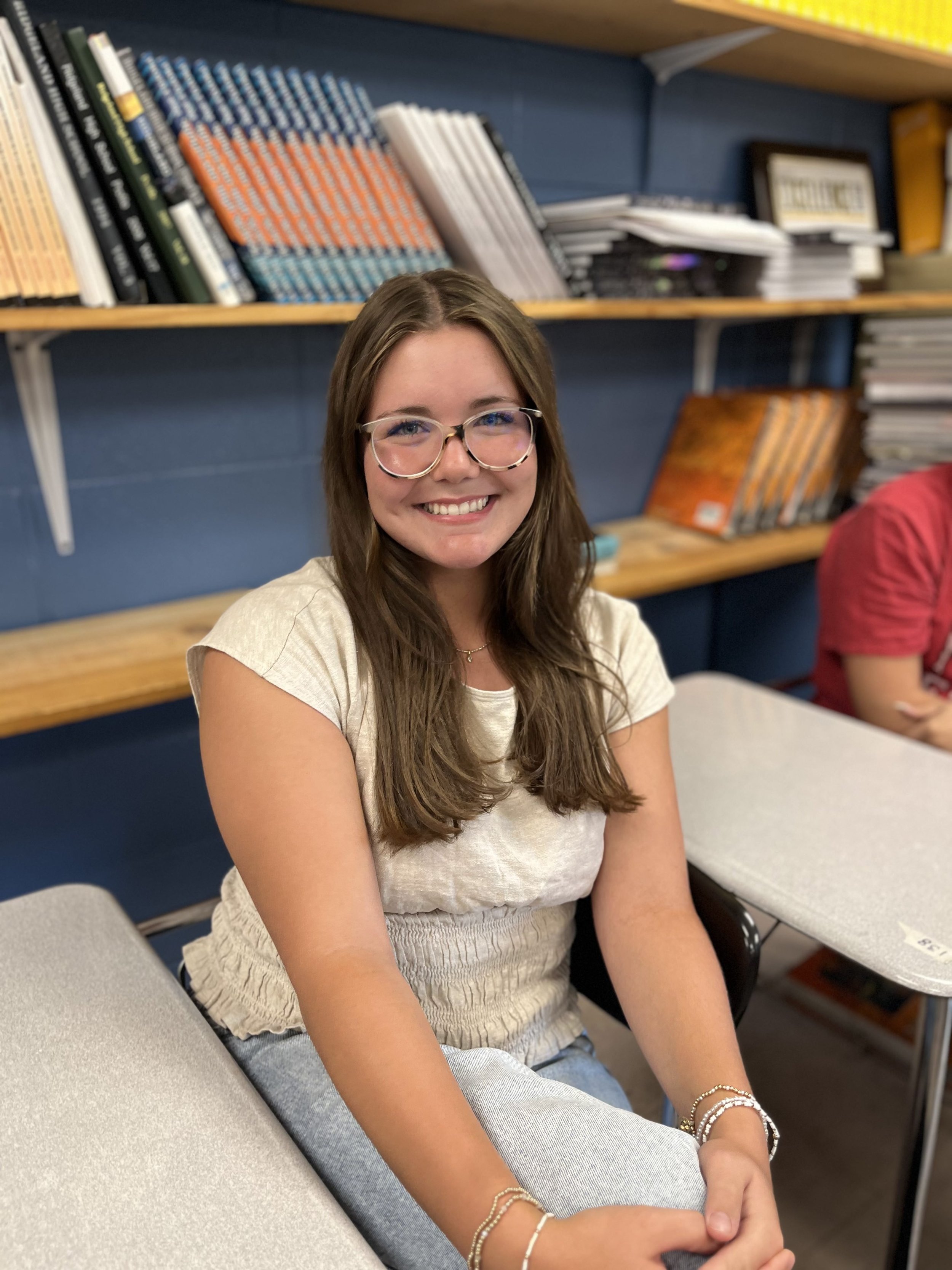 A young woman with glasses smiling, wearing a gray sports jersey with 'Panthers Softball' printed on it, sitting at a table in a classroom with a whiteboard in the background.
