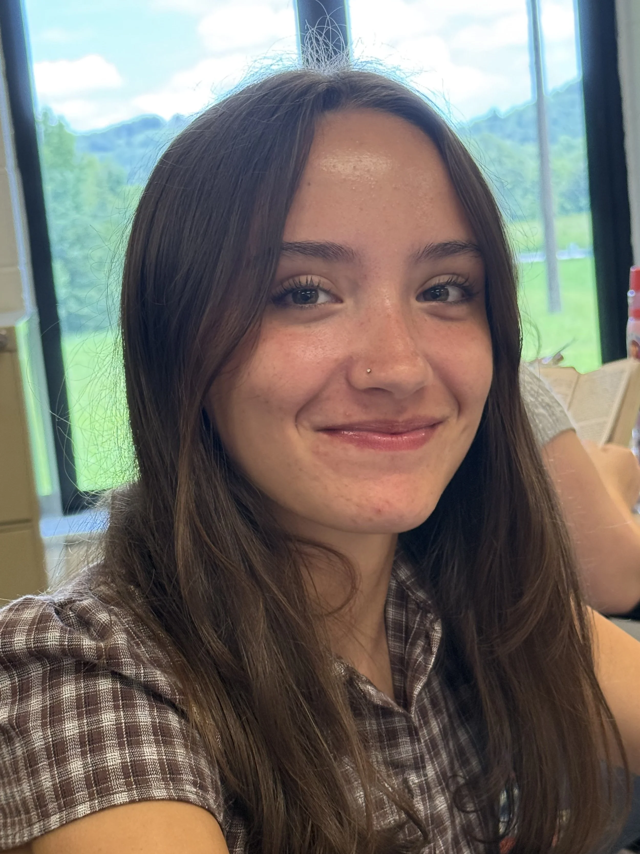 A girl with long brown hair smiling while sitting at a table with laptops in a classroom setting.