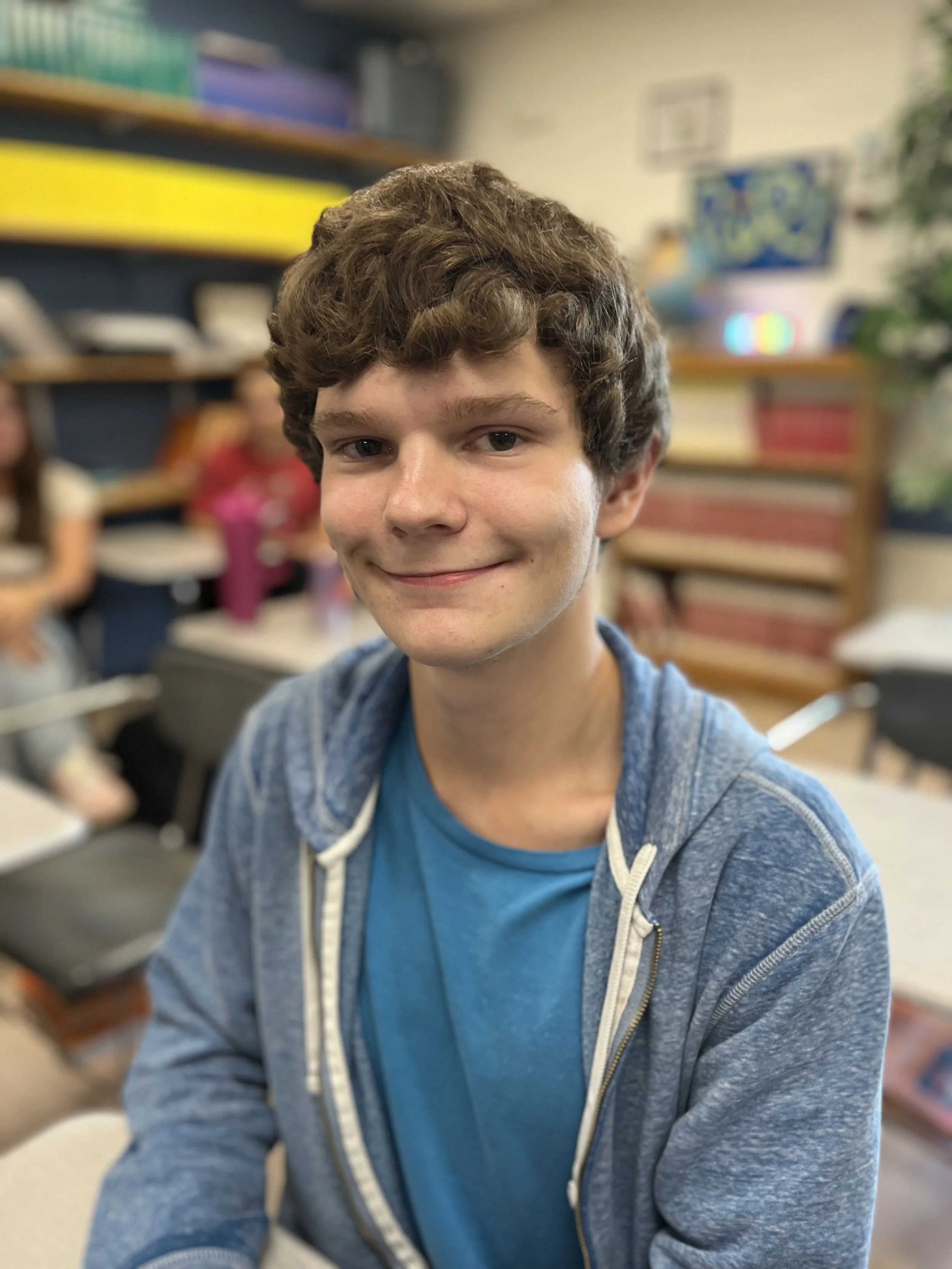 Close-up of a teenage boy smiling, wearing a blue T-shirt and a light blue hoodie, indoors.