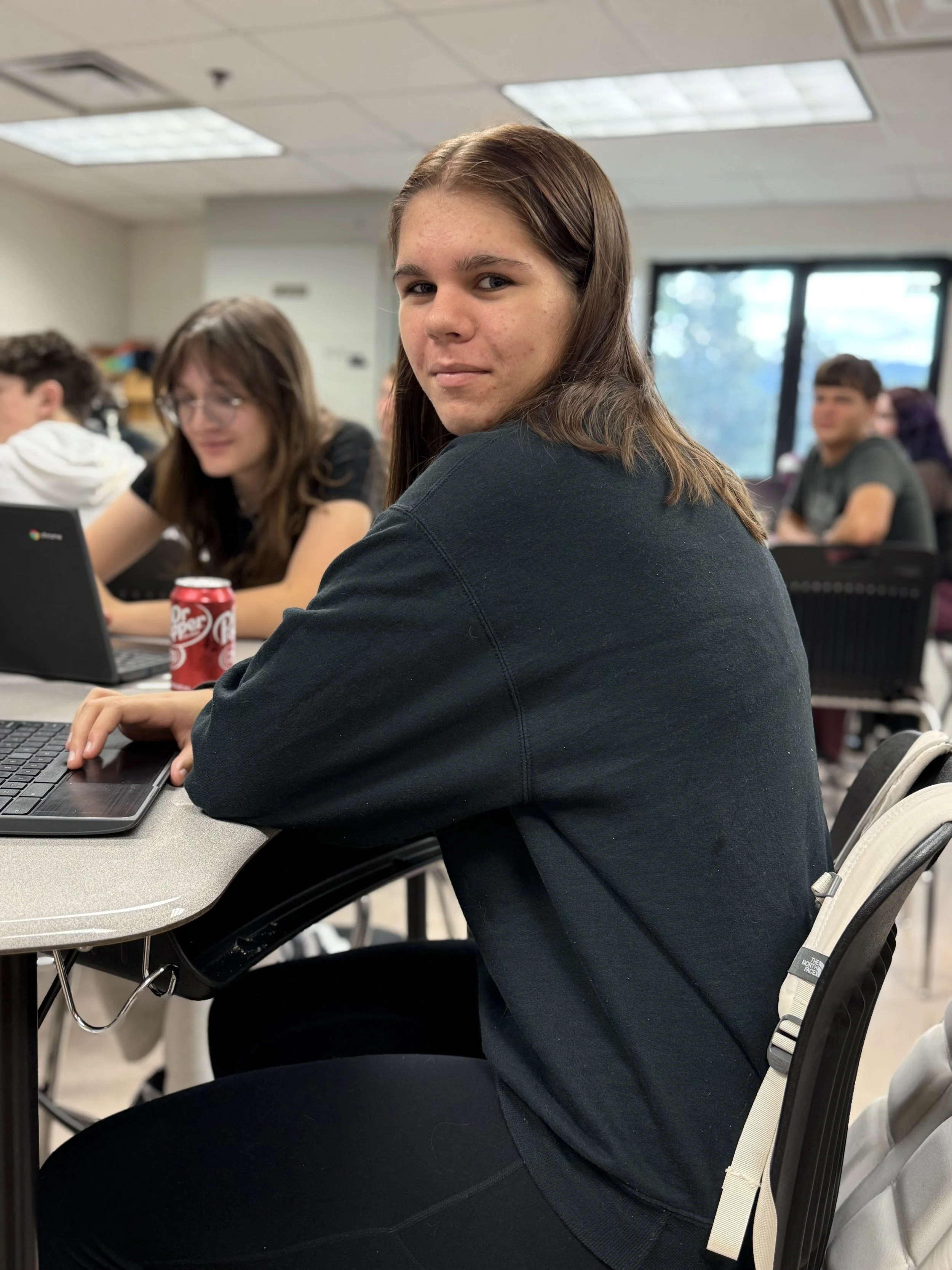 Teen girl with brown hair and a black sweatshirt sitting at a school desk in a classroom, looking at the camera, with other students working on laptops around her.