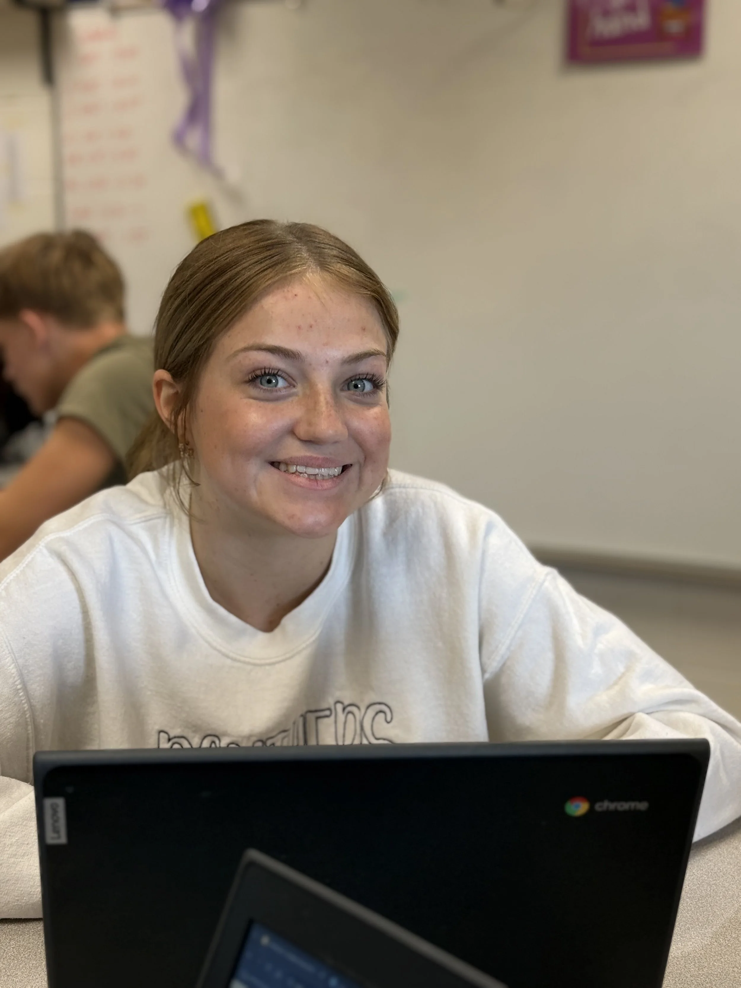 Smiling young woman with blue eyes sitting at a desk with a Chromebook in front of her, in a classroom setting.