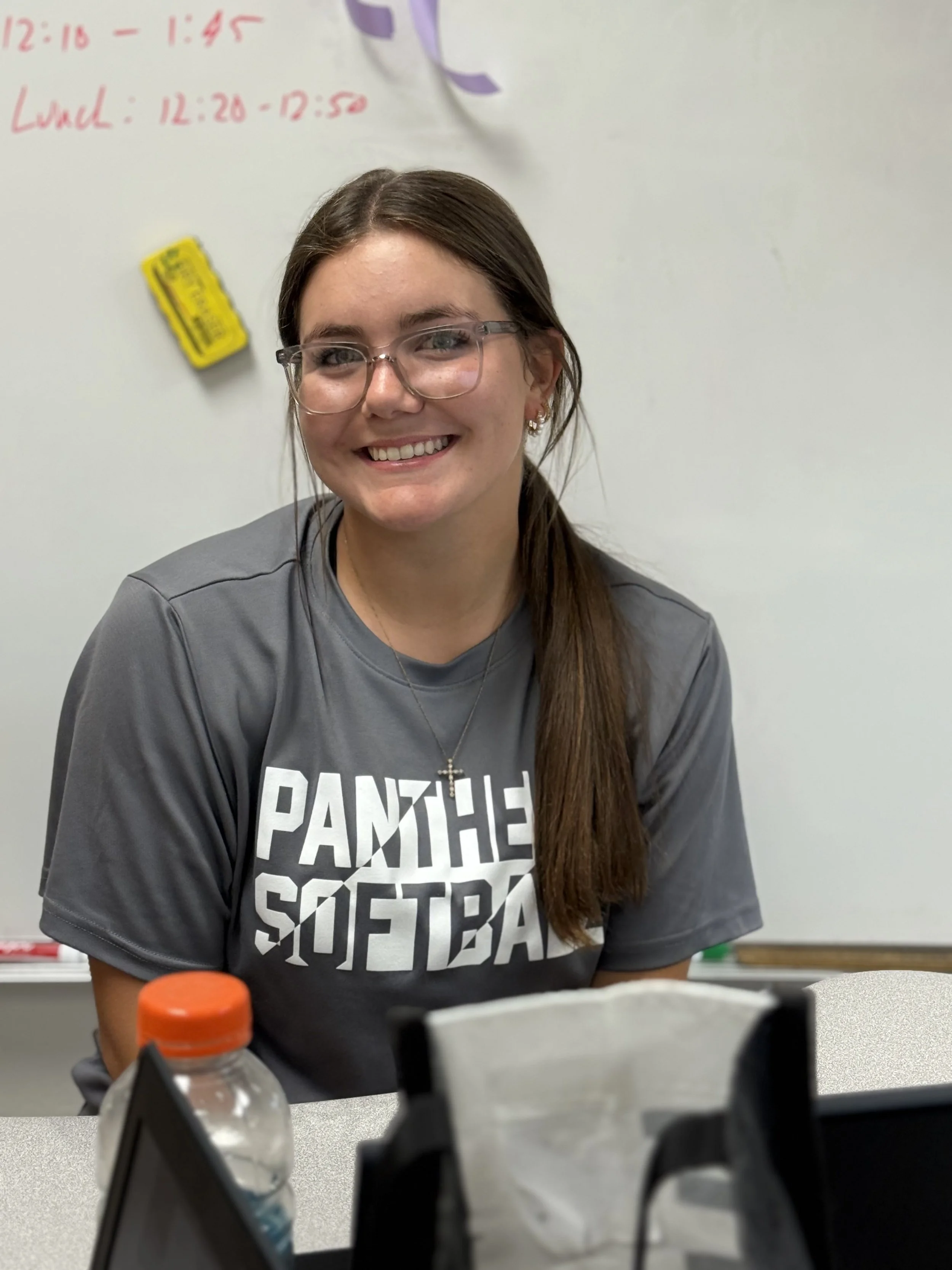 A young woman with glasses smiling, wearing a gray sports jersey with 'Panthers Softball' printed on it, sitting at a table in a classroom with a whiteboard in the background.