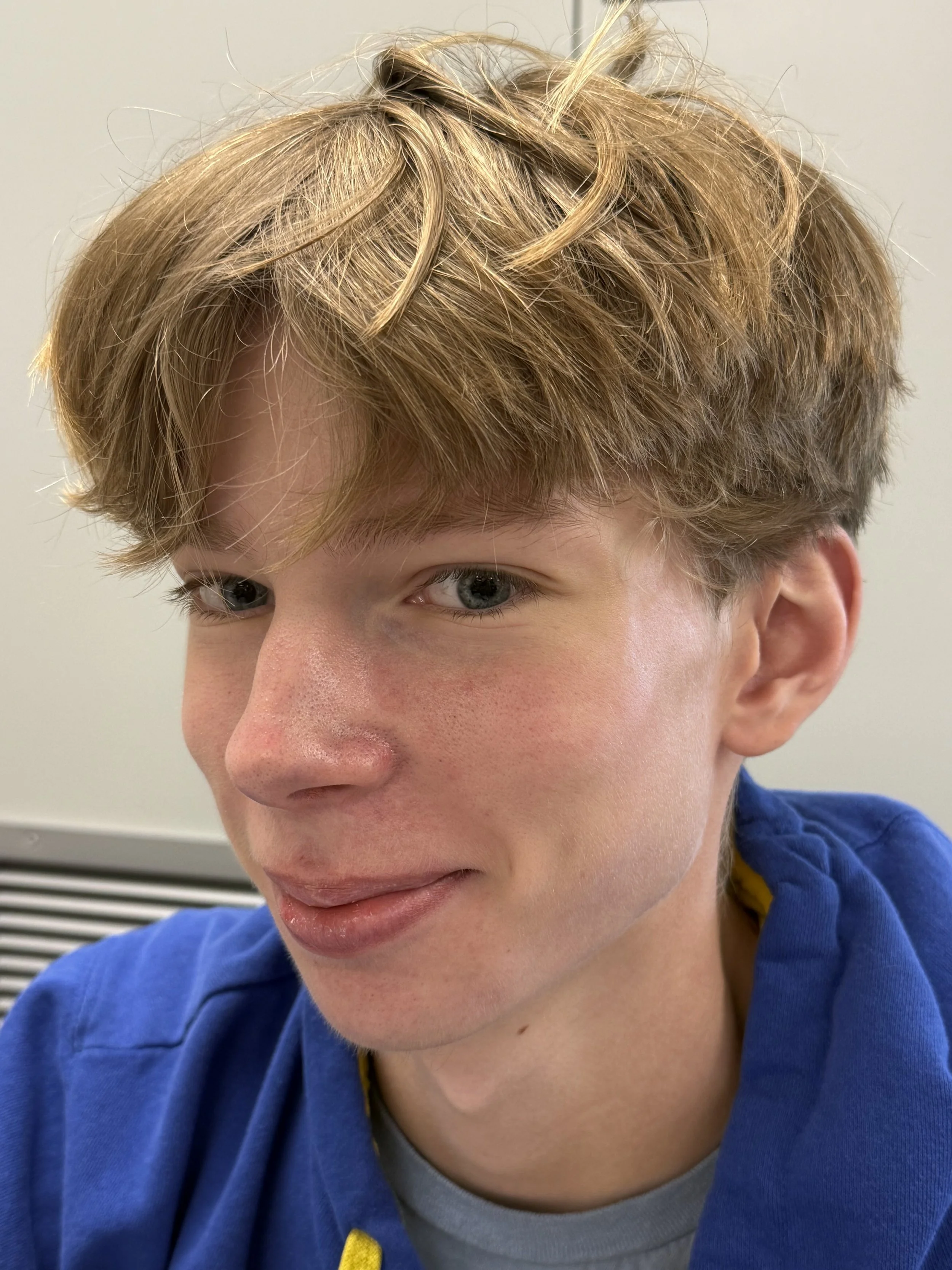 A teenage boy with blond hair sitting on a windowsill, holding a Chromebook in his lap, wearing a purple sweatshirt that says 'DESTIN FLORIDA 1845,' looking at the camera with a slight smile.