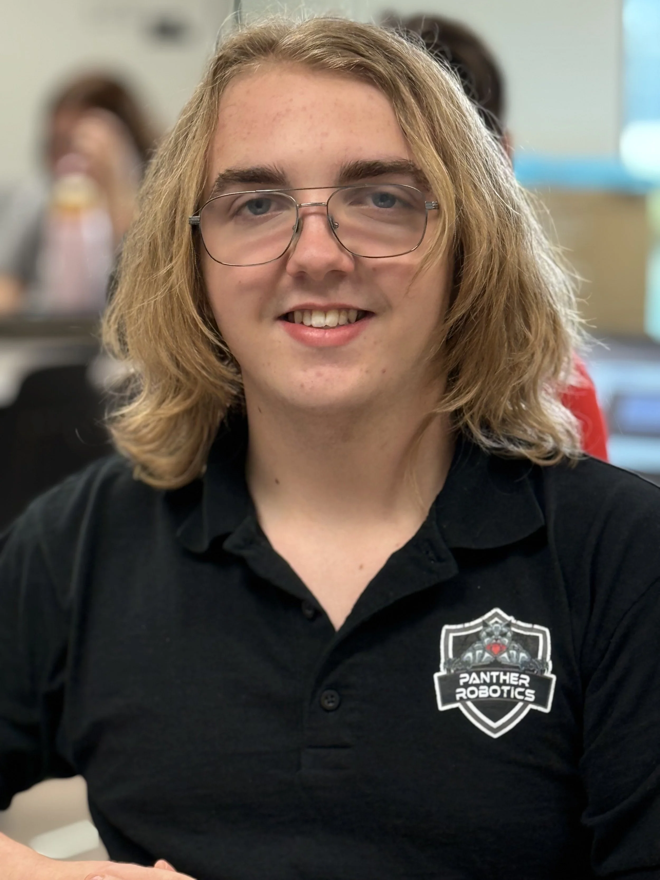 Young man with shoulder-length blond hair, glasses, wearing a black polo shirt with a 'Panther Robotics' logo, smiling at the camera in an indoor setting.