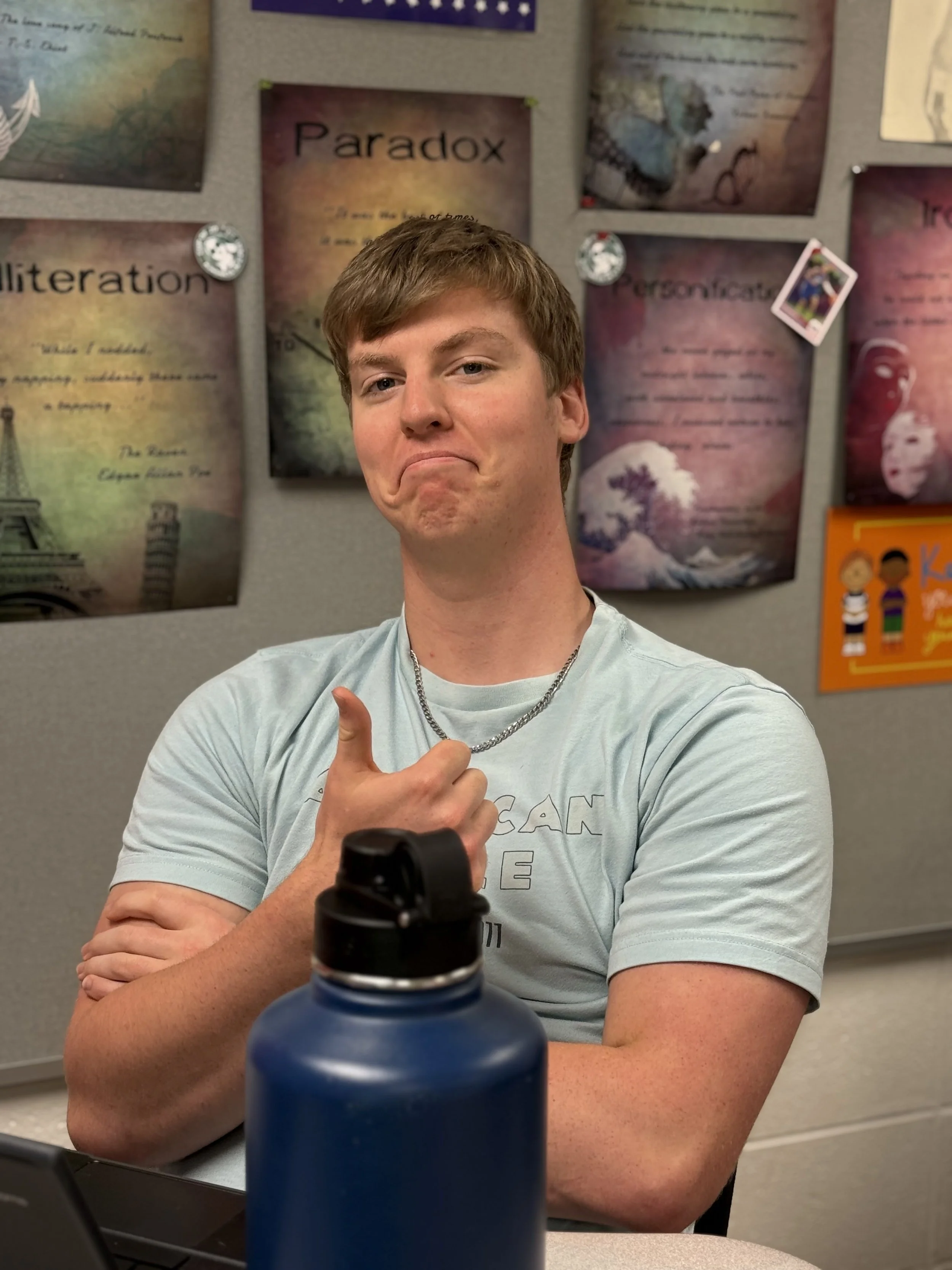 A young man with light brown hair making a sassy face, pointing his finger at himself, seated at a table with a blue water bottle in front of him, in a classroom with posters on the wall behind him.