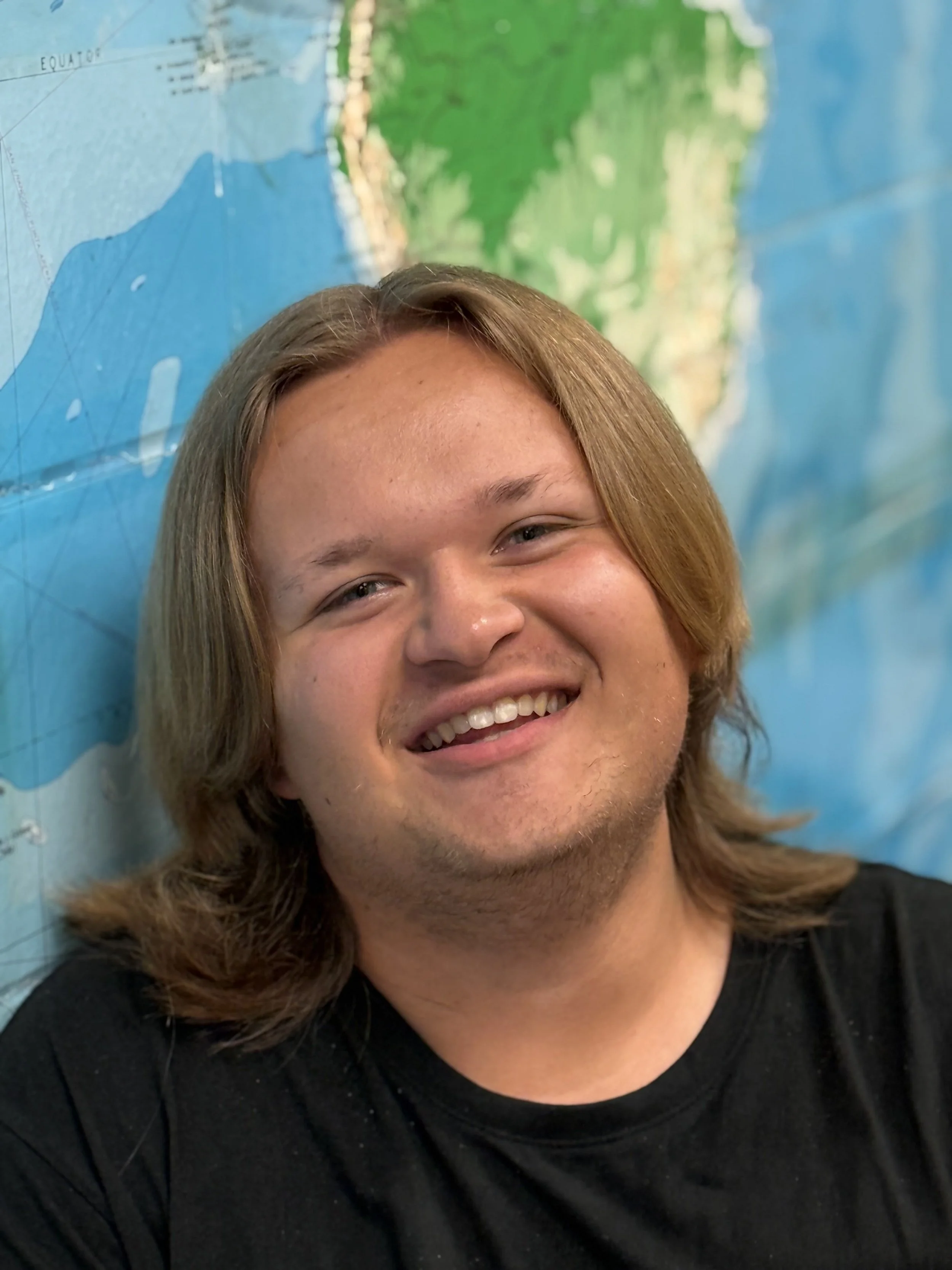 Young man with shoulder-length light brown hair smiling in front of a world map.