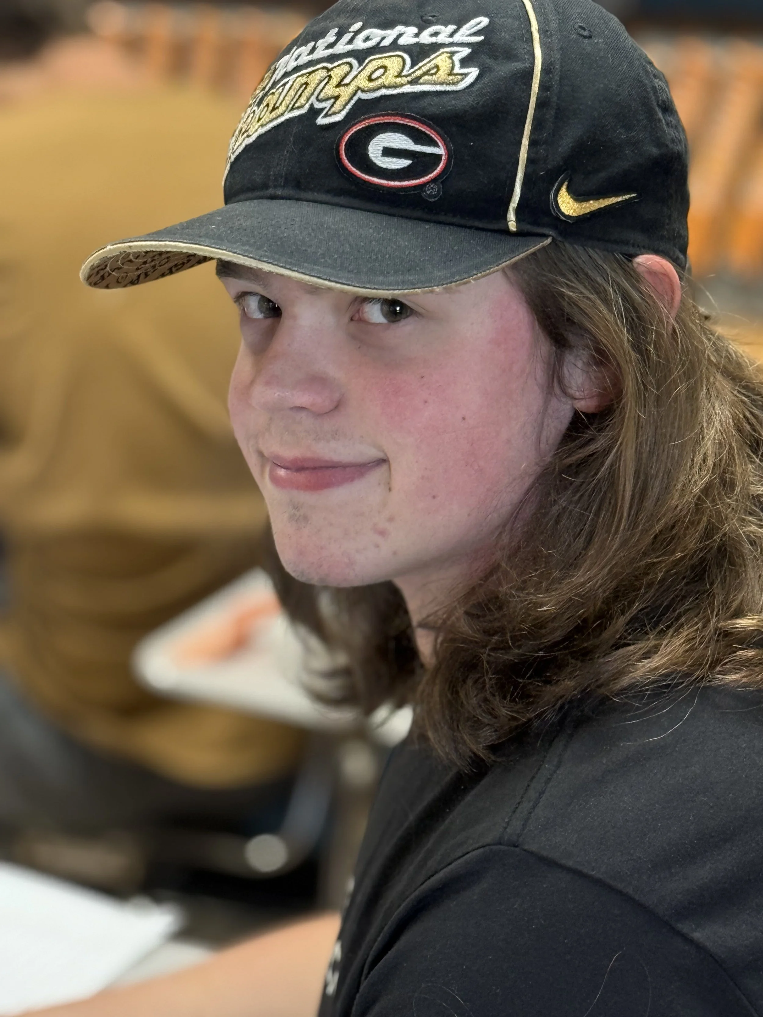 Young man with long brown hair, wearing a Georgia Bulldogs cap with 'National Champions' embroidered on it and a Nike logo, smiling at the camera in an indoor setting.