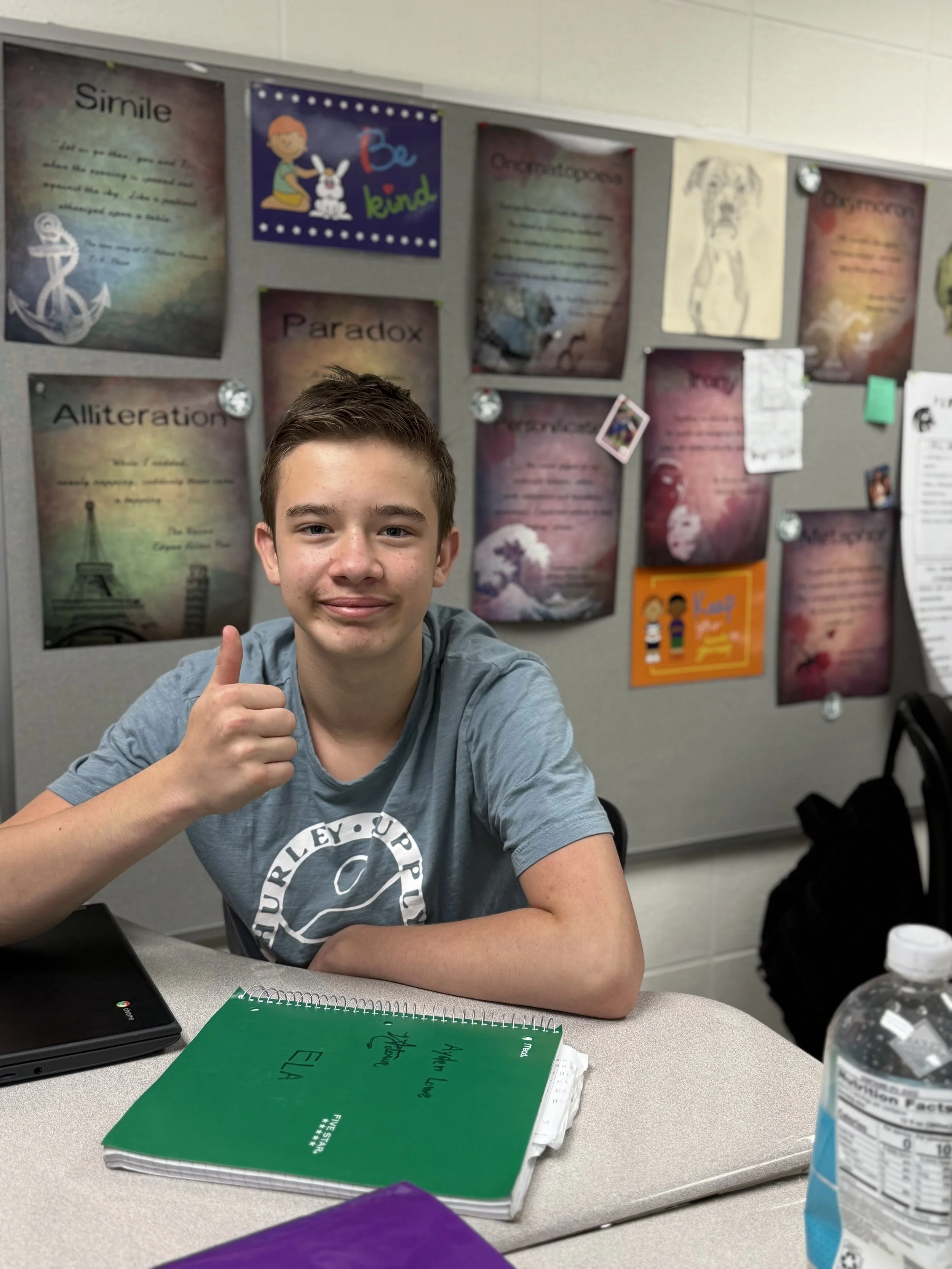 A young boy sitting at a table giving a thumbs-up, with a classroom bulletin board behind him. The bulletin board has various colorful posters and papers, including one with a rainbow and the words "Be kind" and others with text about different topics. The table has a green notebook, a laptop, and a bottle of water.