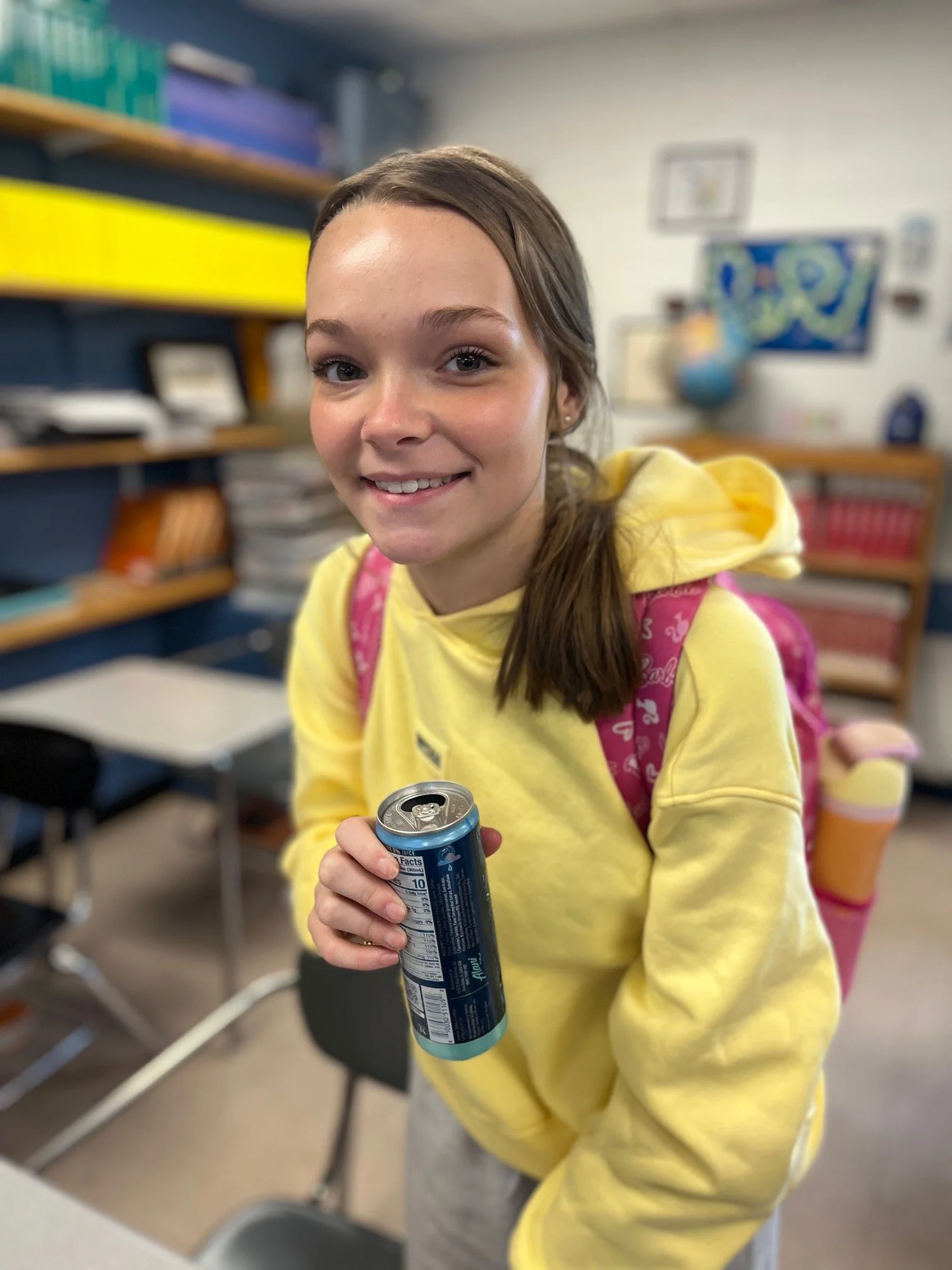 A teenage girl with light brown hair in a bun, wearing a gray hoodie, sitting at a desk with a notebook and a blue pen, smiling slightly at the camera in a classroom setting.