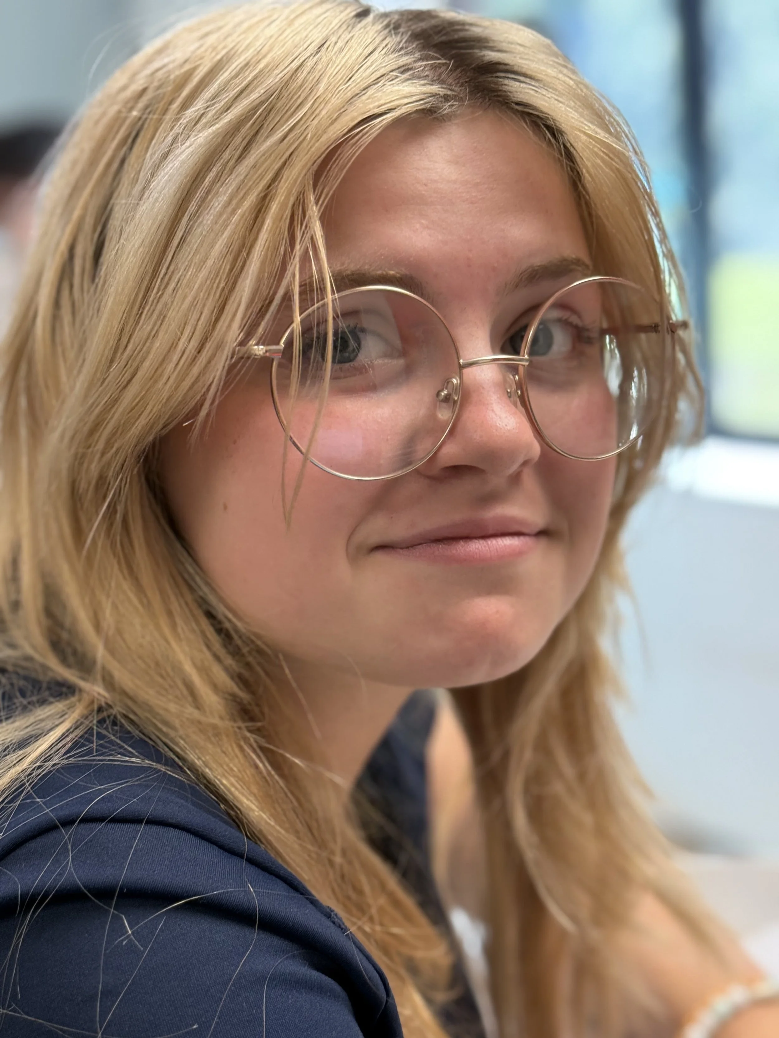 A young woman with reddish-blonde hair wearing round glasses and a dark jacket, smiling subtly, indoors with a blurred background.
