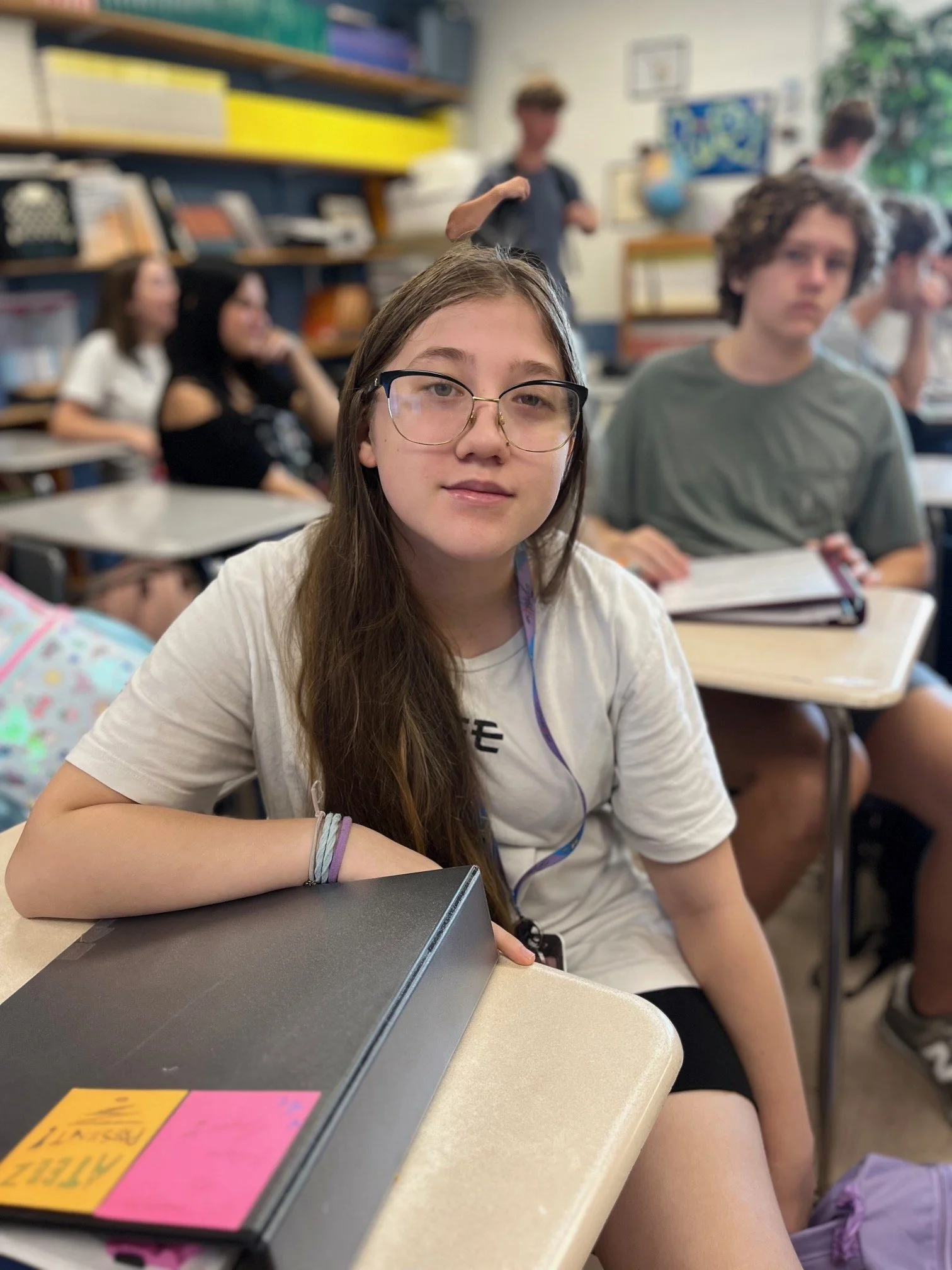 A teenage girl with long, curly, dark red hair, smiling at the camera, sitting at a desk in a classroom with other students in the background.