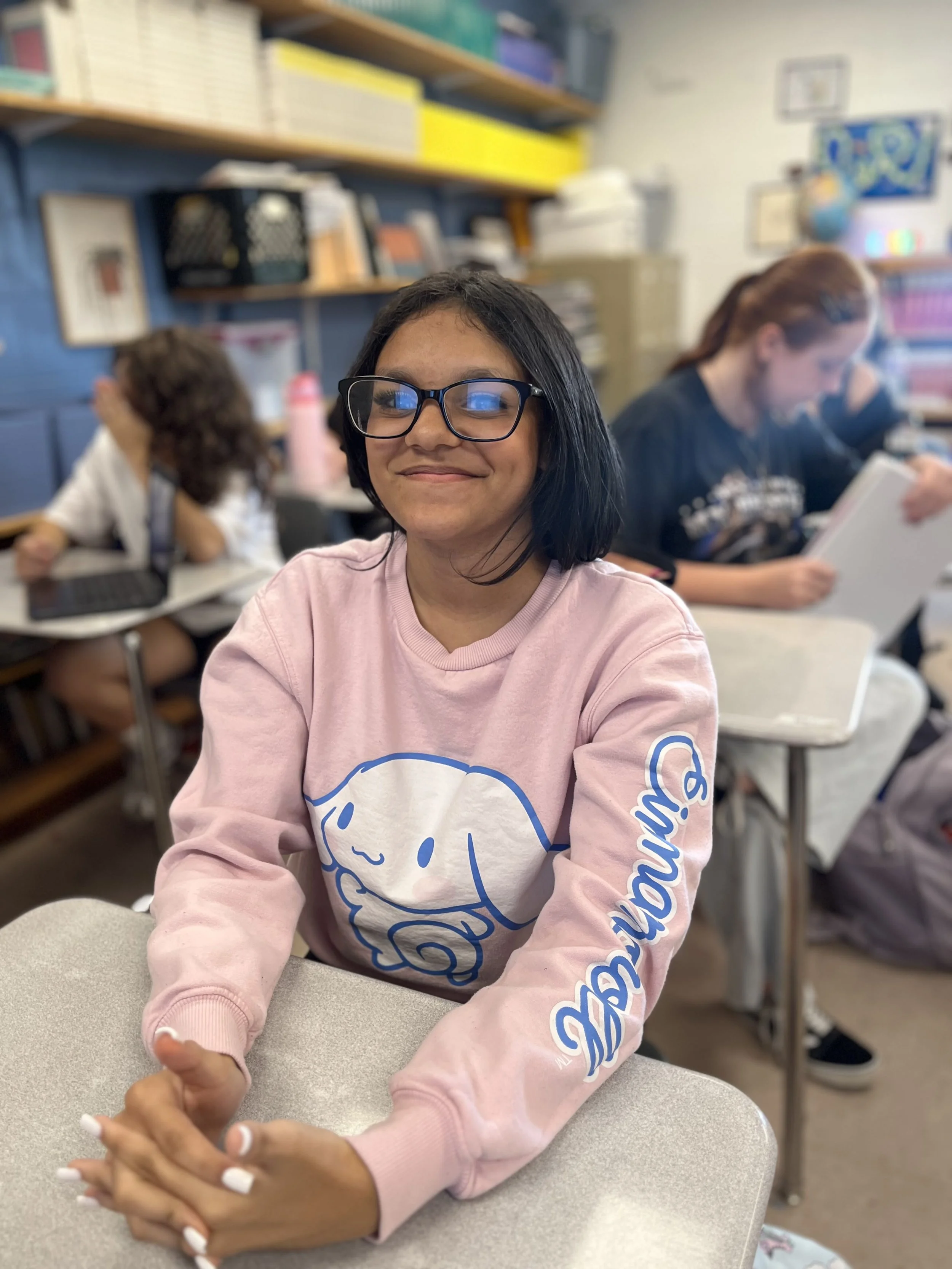 A young girl wearing glasses and a red shirt sitting at a table, resting her chin on her hand, looking directly at the camera. She has a smartwatch on her wrist and is in a room with other people in the background.