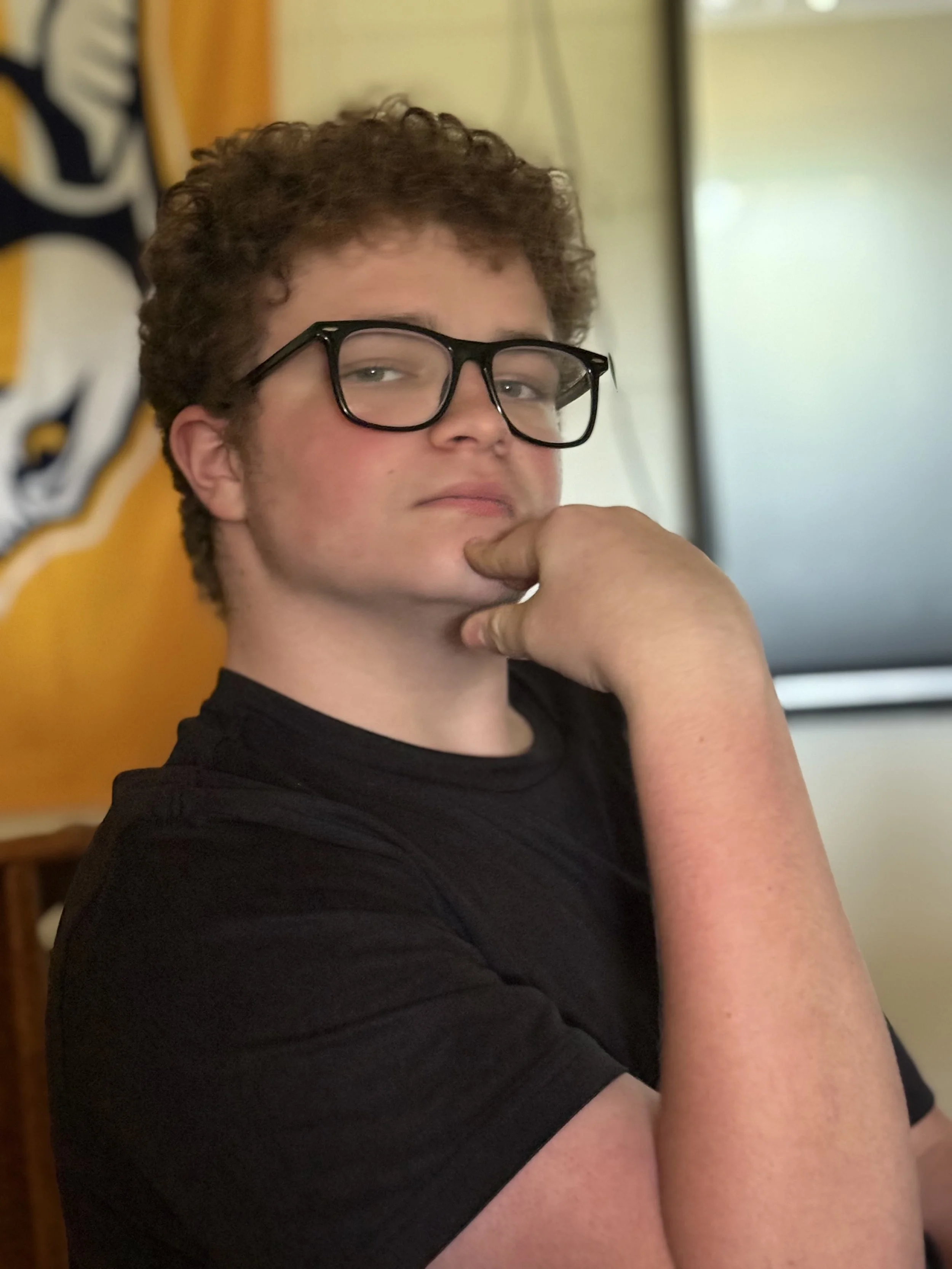 A young person with curly hair and glasses resting their chin on their hand, looking at the camera.