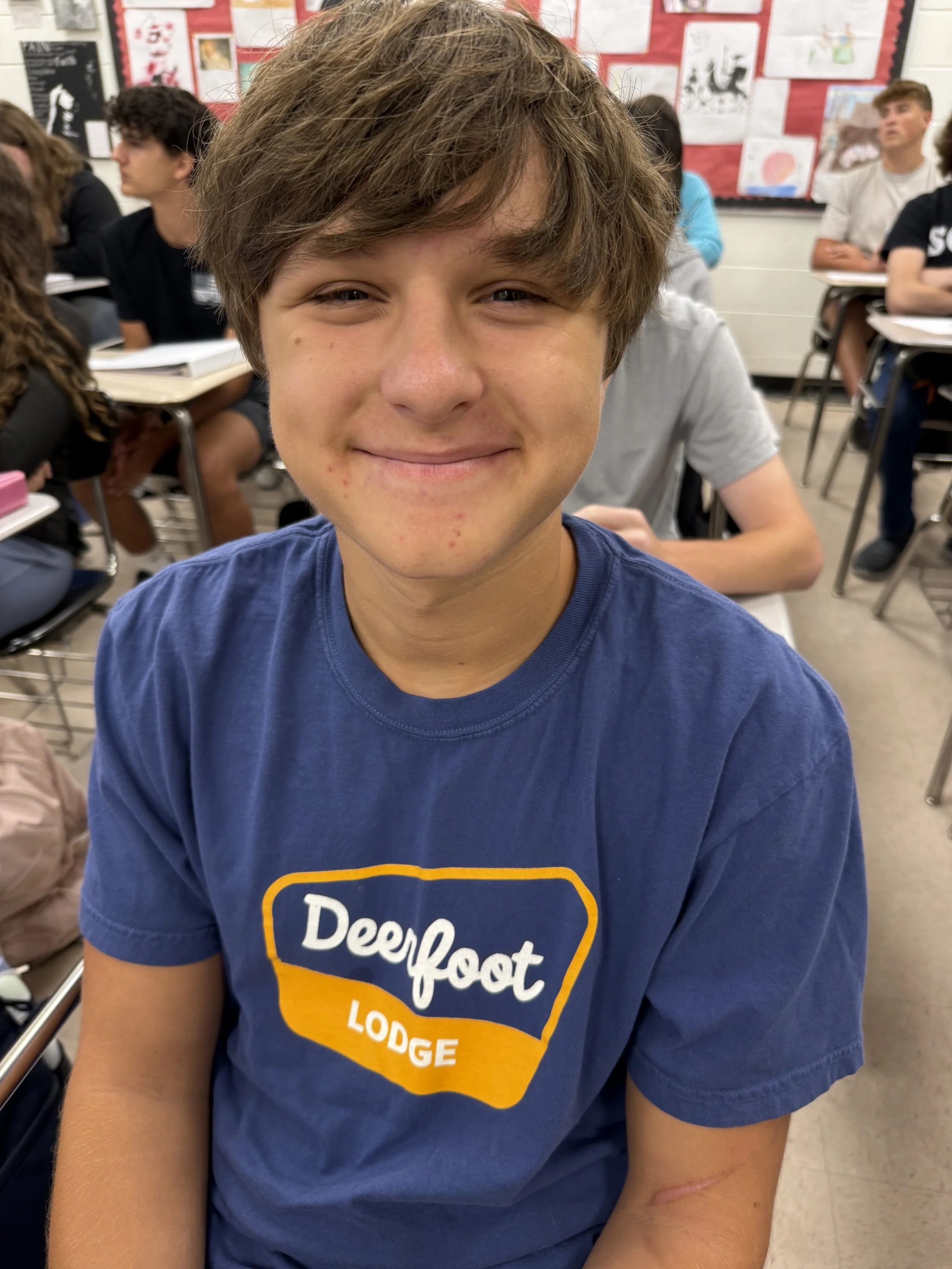 A teenage boy with light skin, brown hair, and a slight smile, wearing a blue T-shirt with white text and graphics, standing indoors.