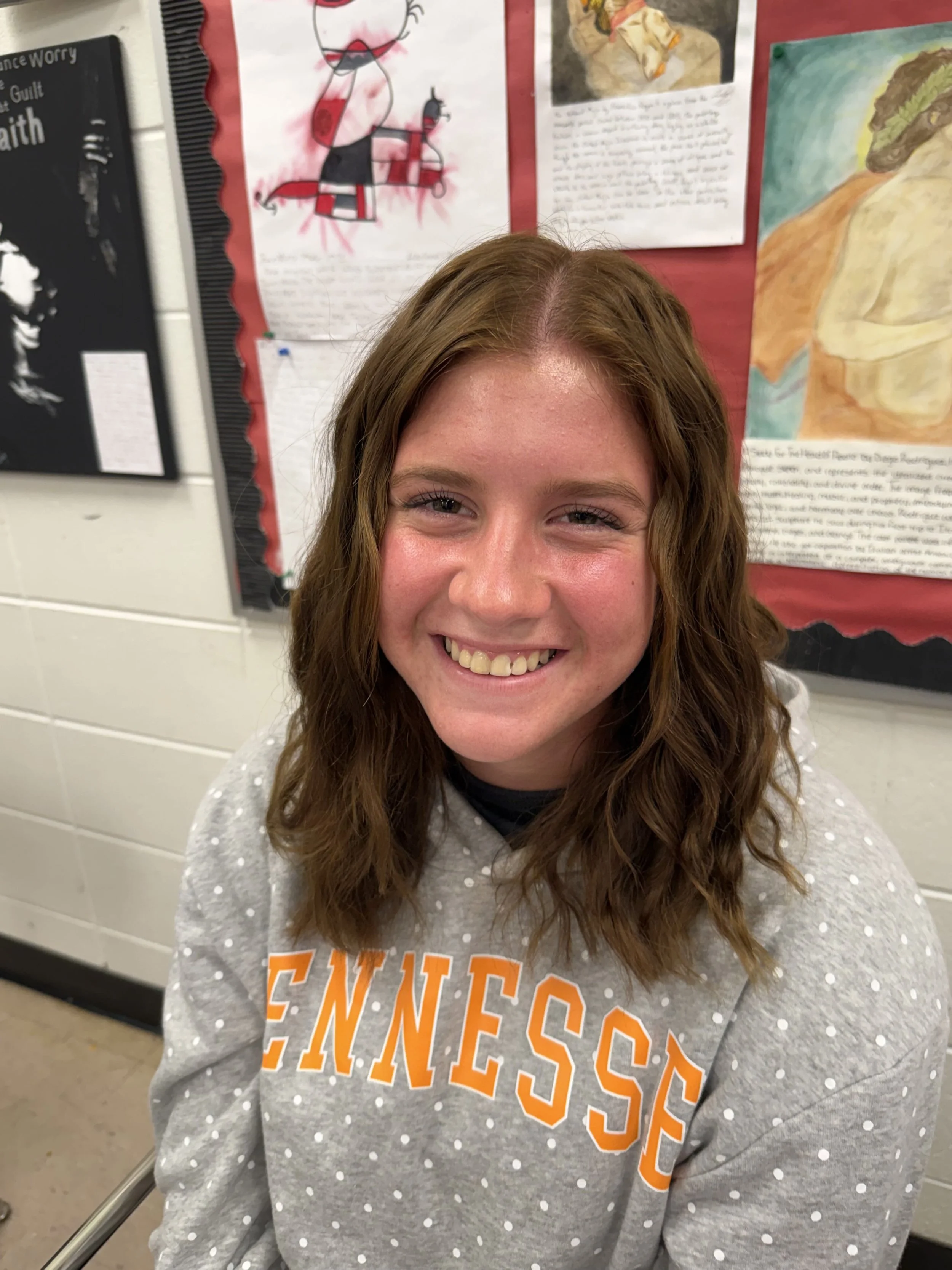 Close-up of a smiling young woman with red hair, braces, and bright eyes, wearing a light pink shirt.