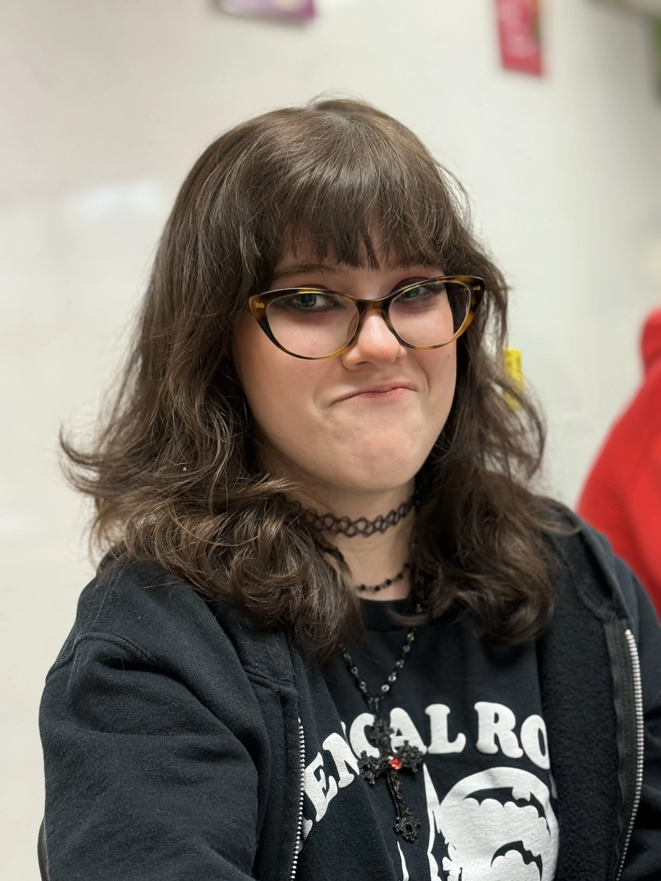 A young woman with brown, curly hair and glasses, wearing a black shirt with white text and a black hoodie, and jewelry including a choker and beaded necklace with a cross pendant, smiling slightly.