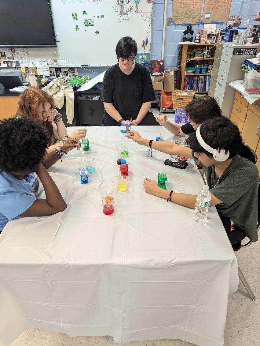 A group of five young people sitting around a white table, playing a game with small colored objects, in a classroom or office setting.