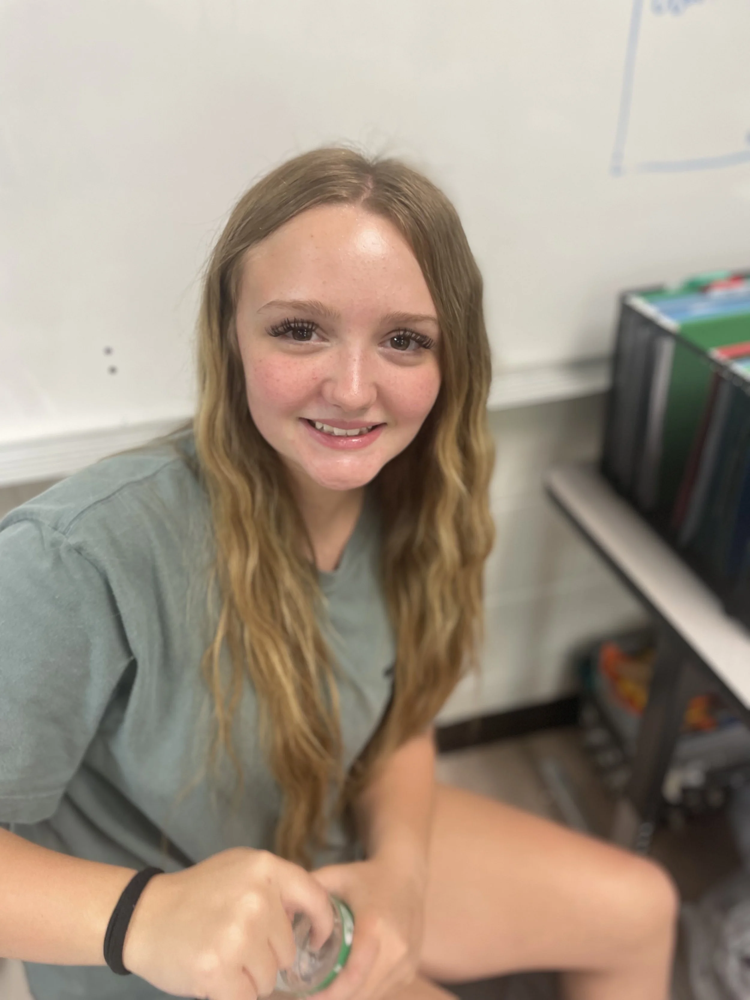 A young woman with long wavy hair and freckles, smiling at the camera, sitting in a classroom with a whiteboard and shelves of binders behind her.