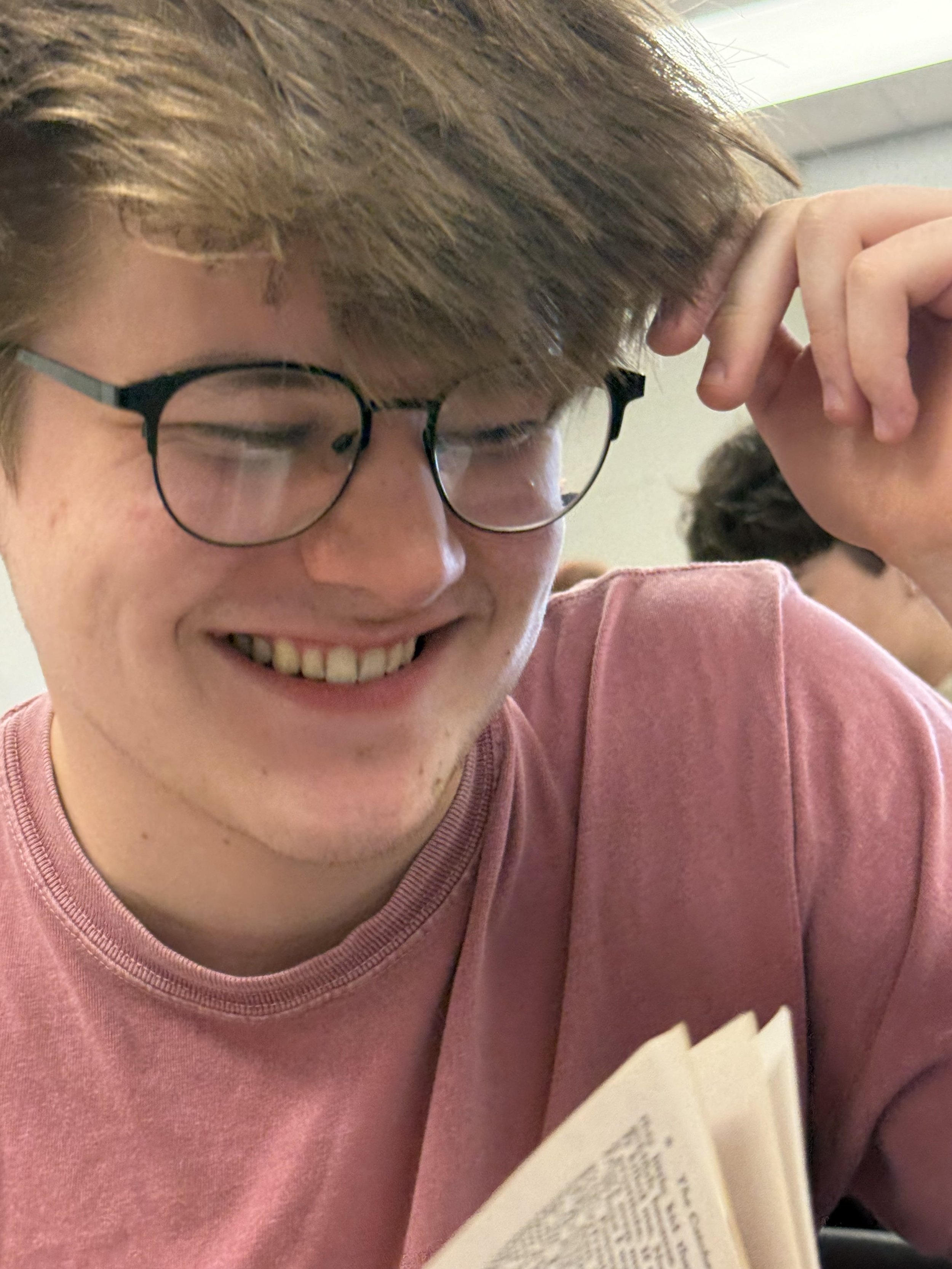A young man with glasses and tousled brown hair sits at a desk in a classroom, wearing a black hoodie, with a notebook and laptop in front of him.