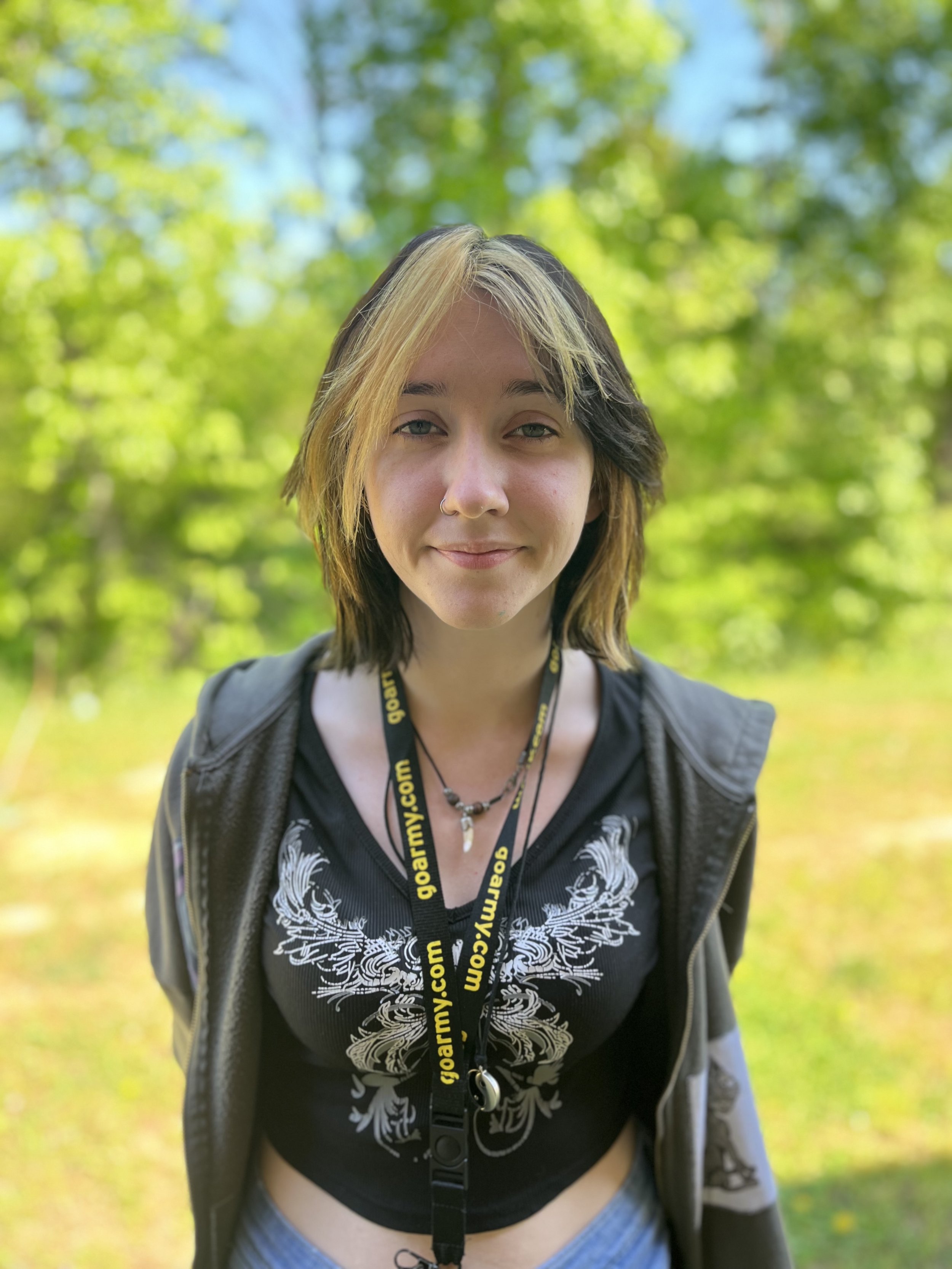 A young woman with glasses and long dark hair sitting at a desk, smiling at the camera. She wears a black top, gray cardigan, and a necklace. The background includes blue walls, shelves with storage bins, and a framed artwork.